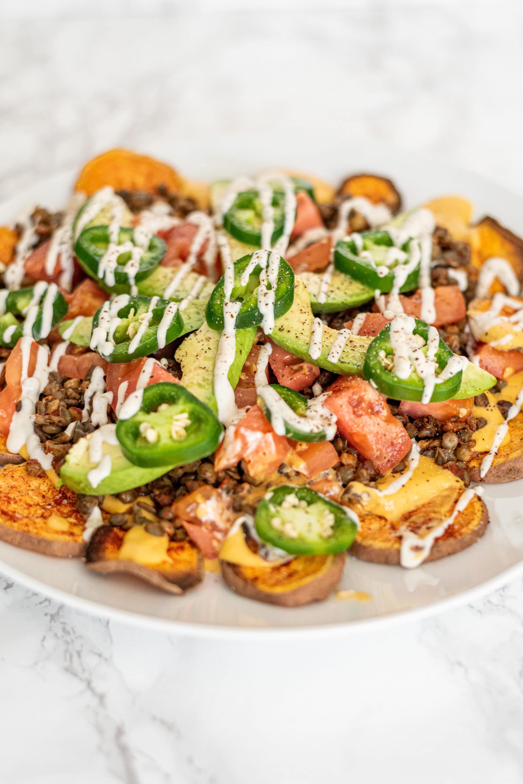 A white plate with sweet potato nachos on it topped with lentil meat, avocado slices, tomato slices, jalapeno slices, and a drizzle of sour cream.