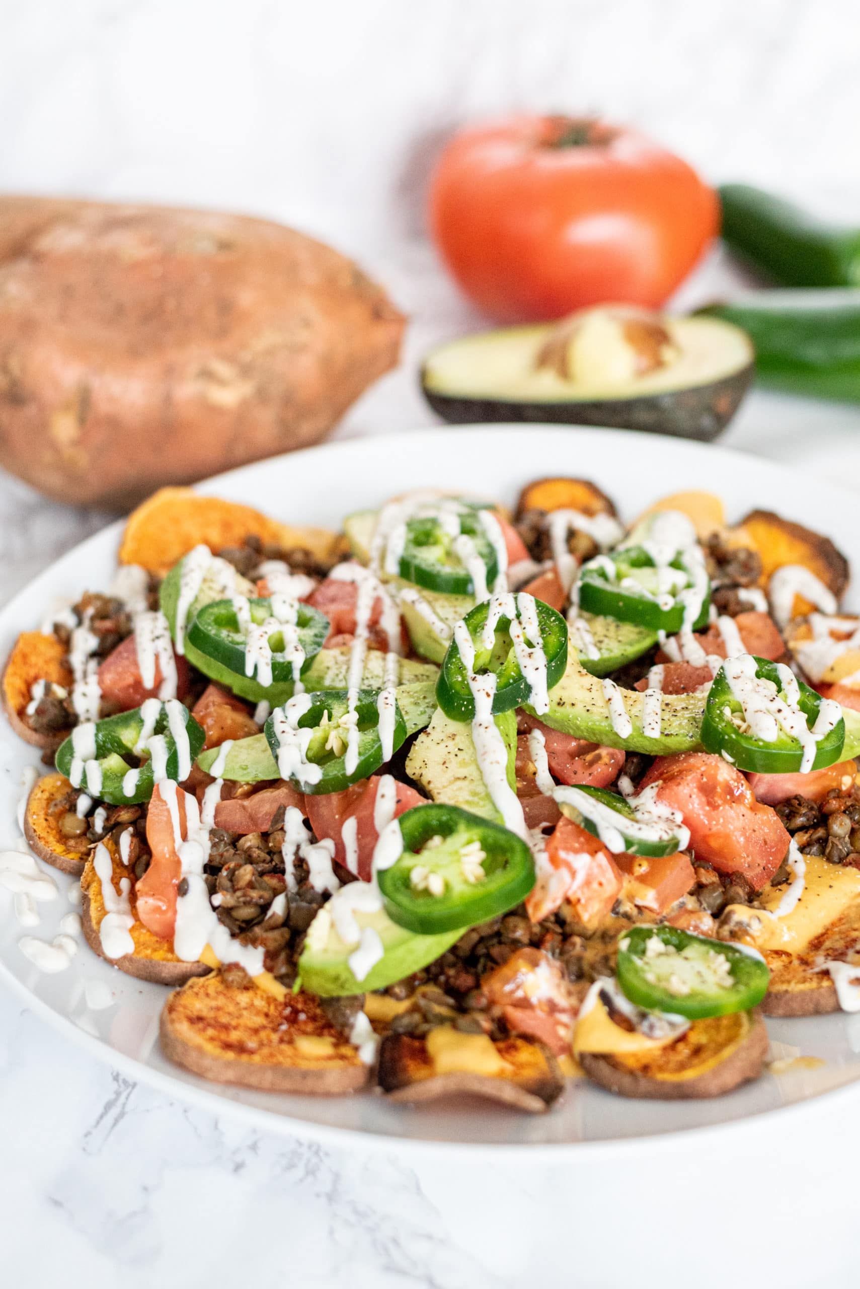 A white plate with sweet potato nachos on it topped with lentil meat, avocado slices, tomato slices, jalapeno slices, and a drizzle of sour cream. Behind the plate is a sweet potato, half an avocado, and a whole tomato.
