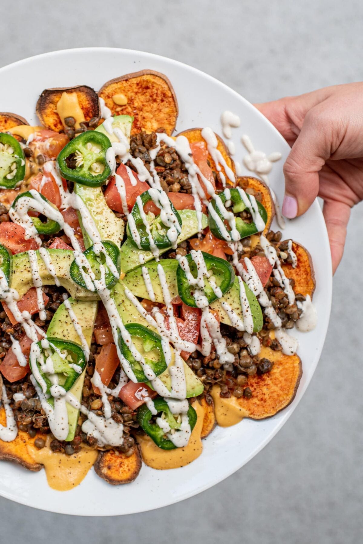 A white plate being held by a white hand with sweet potato nachos on it topped with lentil meat, avocado slices, tomato slices, jalapeno slices, and a drizzle of sour cream.