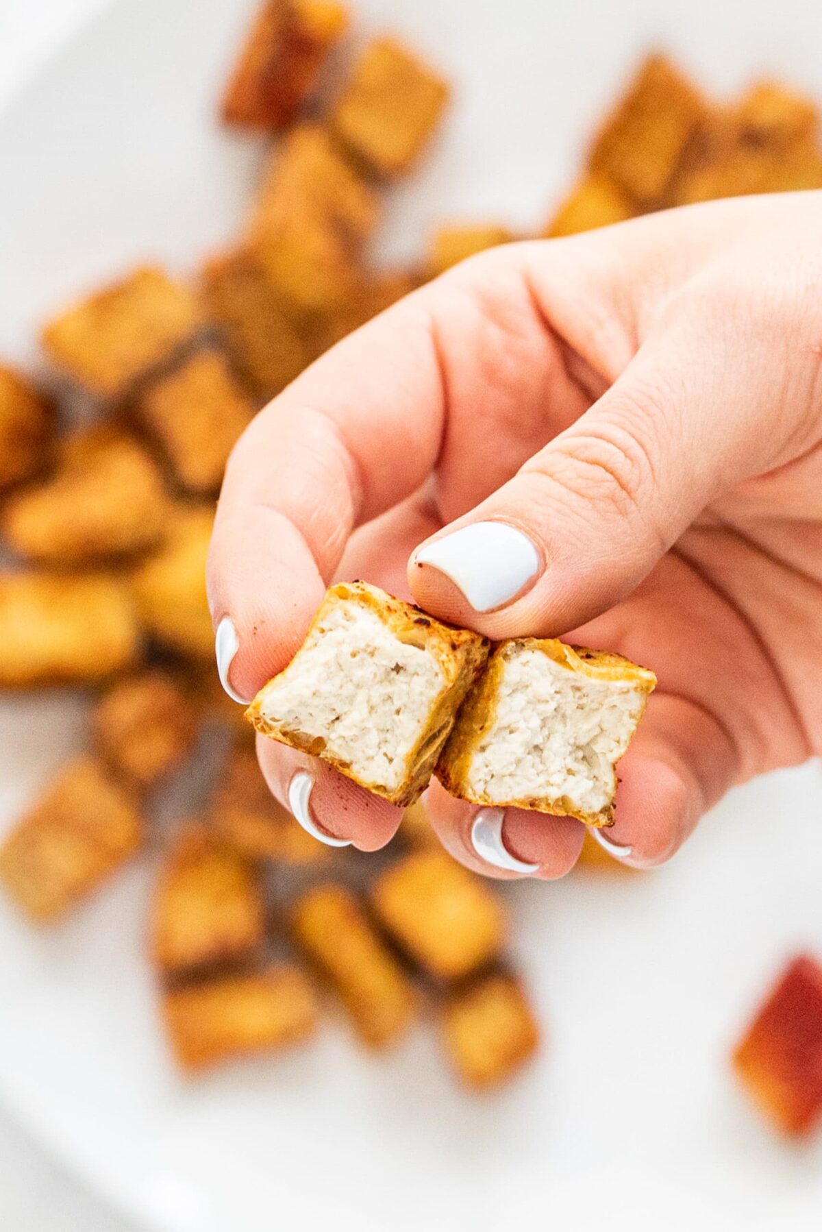 One white hand with painted nails holding a tofu nugget split in half and a white plate in the background of more nuggets.