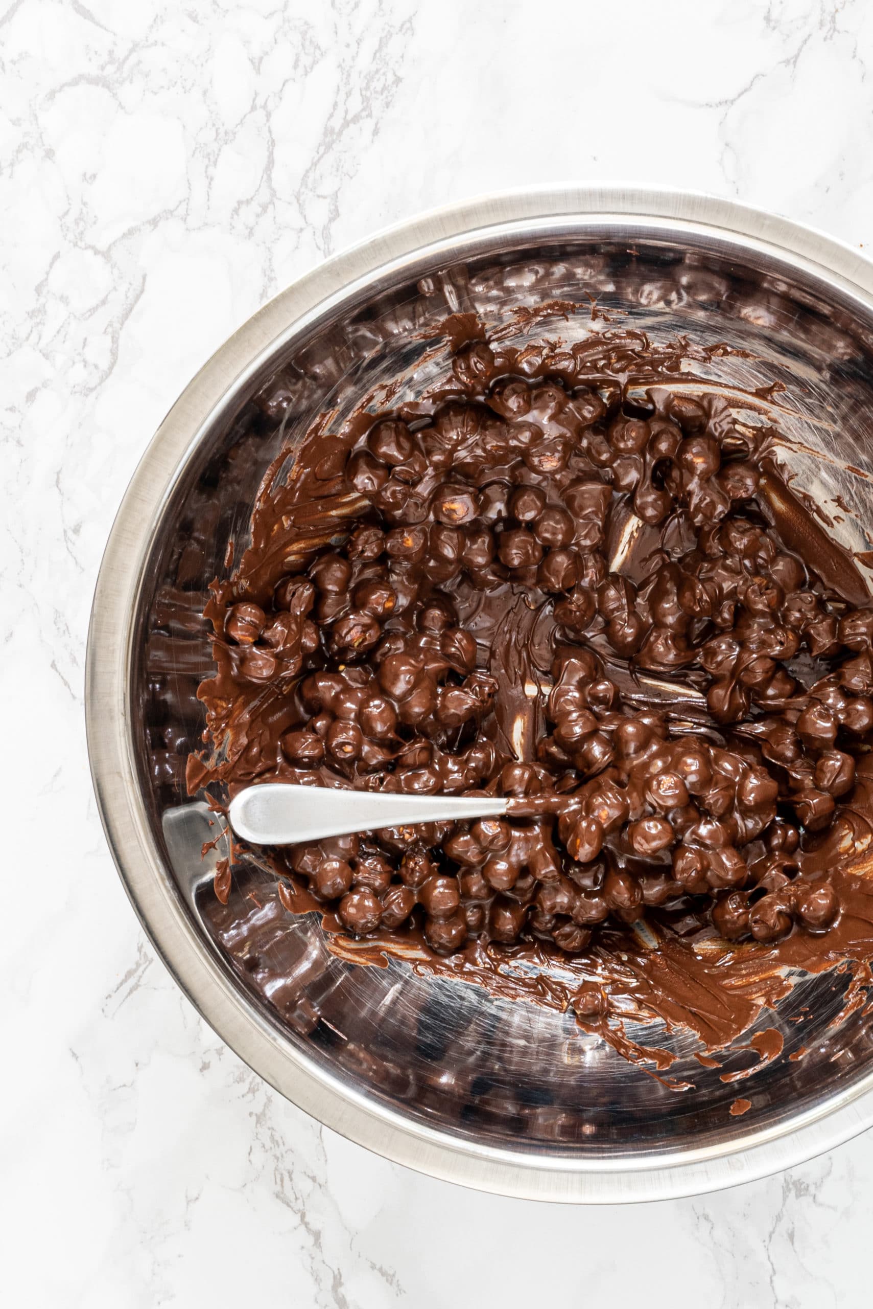 A silver metal bowl filled with chickpeas covered in melted chocolate and a silver spoon sticking out.