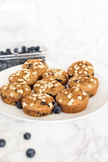 Moist Vegan Blueberry Muffins on a white plate with a carton of blueberries behind them and three blueberries in front of the plate.