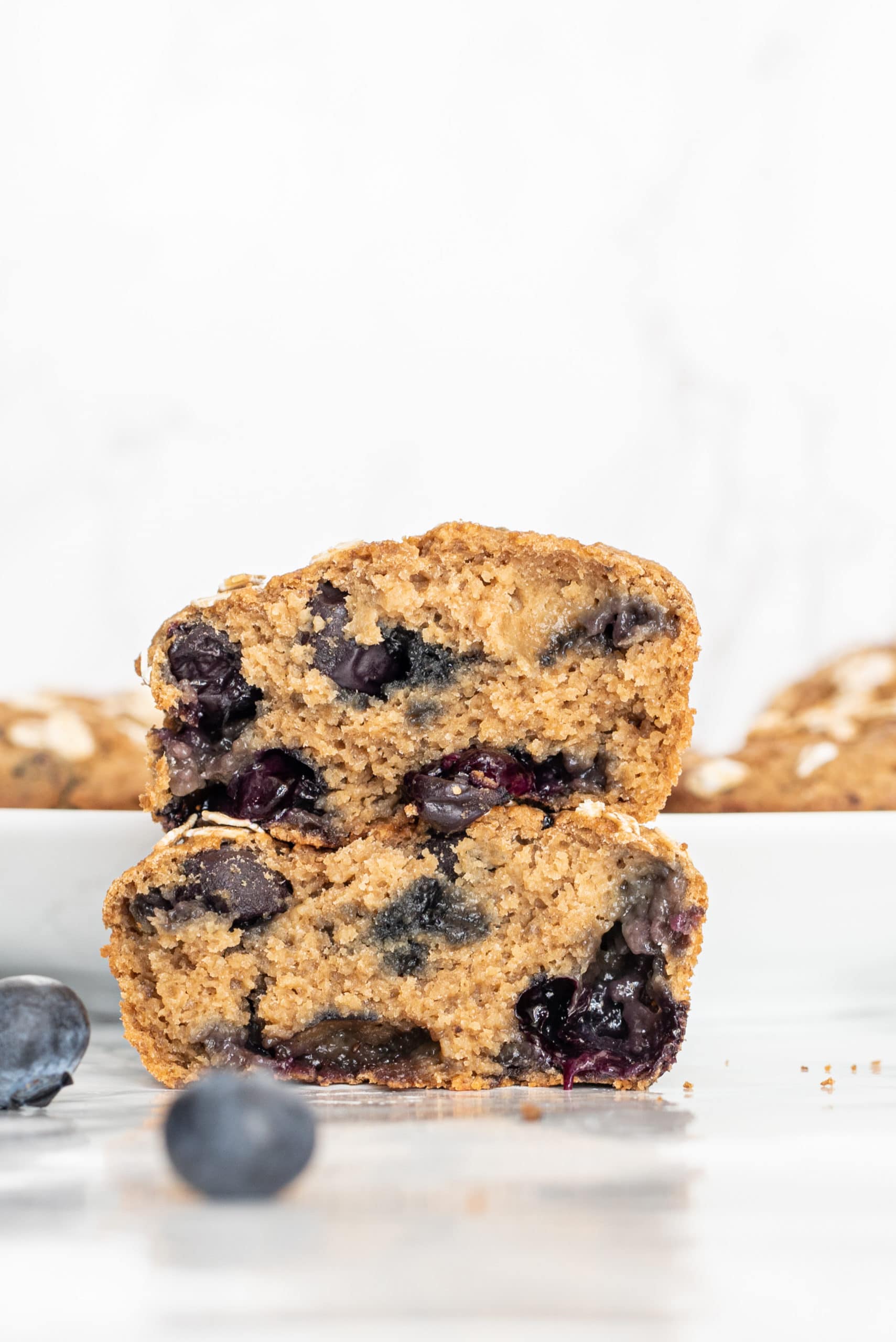 One vegan blueberry muffin split into two halves and stacked on each other to show the inside. There are a couple blueberry muffins in the background and two blueberries in front for decor.