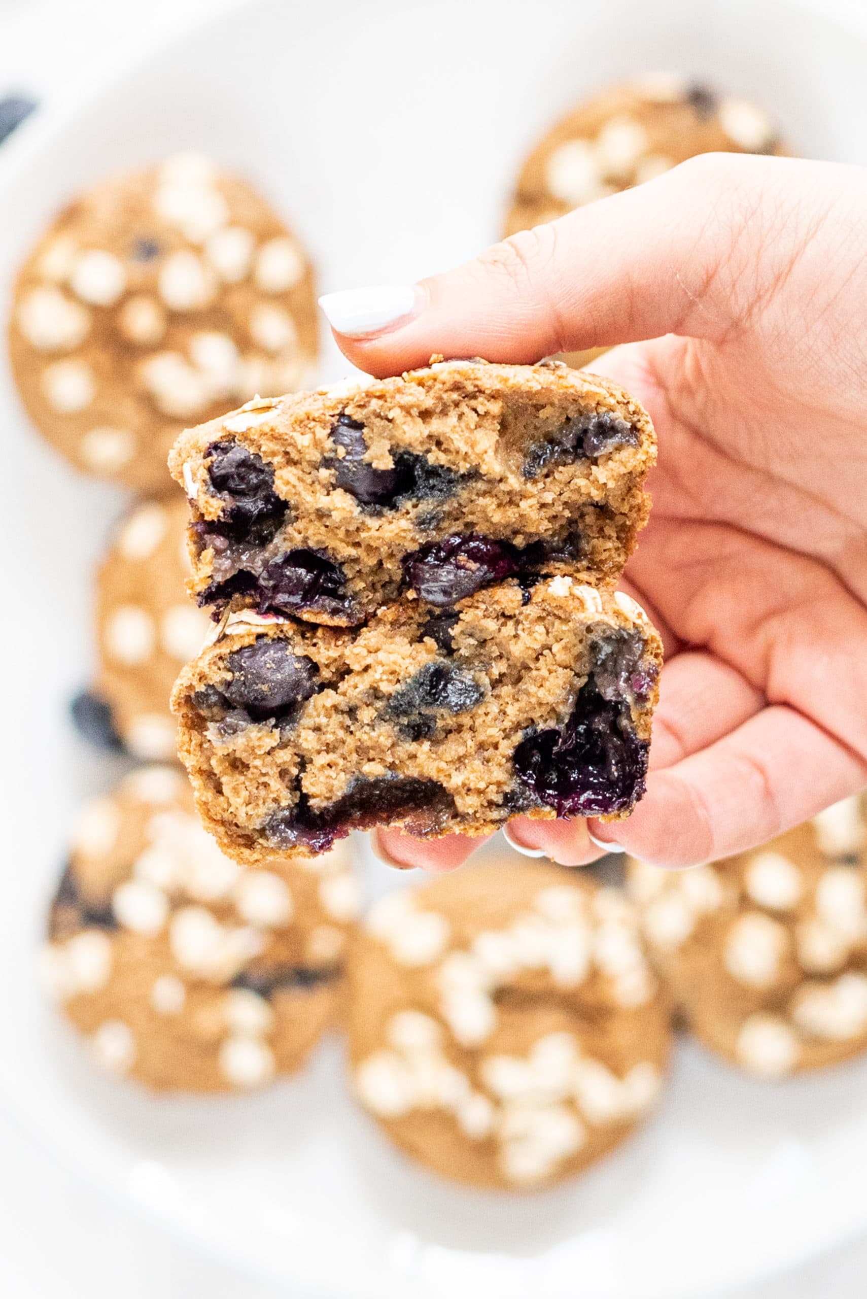 A white plate in the background with blueberry muffins on it. In the center of the photo is a hand with white skin holding a blueberry muffin split in half.