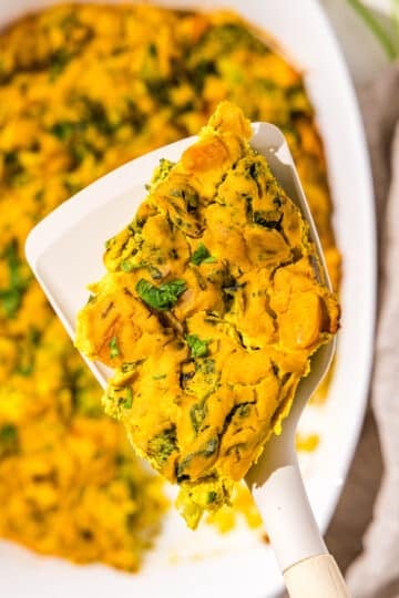 A piece of tofu chickpea frittata on a white spatula overtop a white casserole dish filled with the rest of the frittata.