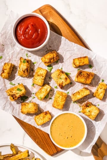 A brown cutting board with a piece of crinkled parchment paper on it topped with air fried tofu nuggets and two bowls of sauce (one is ketchup and one is honey mustard).