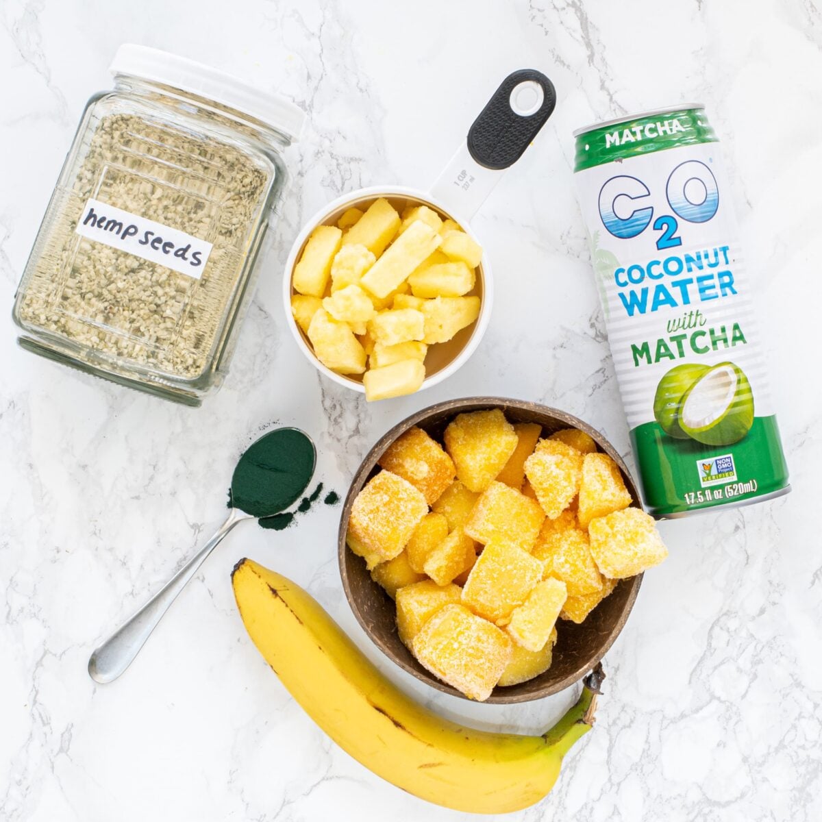 A jar of hemp seeds, a bowl of frozen mango and frozen pineapple, a whole fresh banana, a spoon filled with green spirulina powder, and a bottle of coconut water.