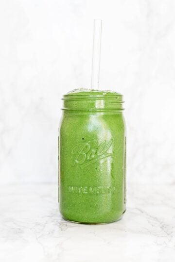 A tall mason jar filled with green smoothie and a glass straw.