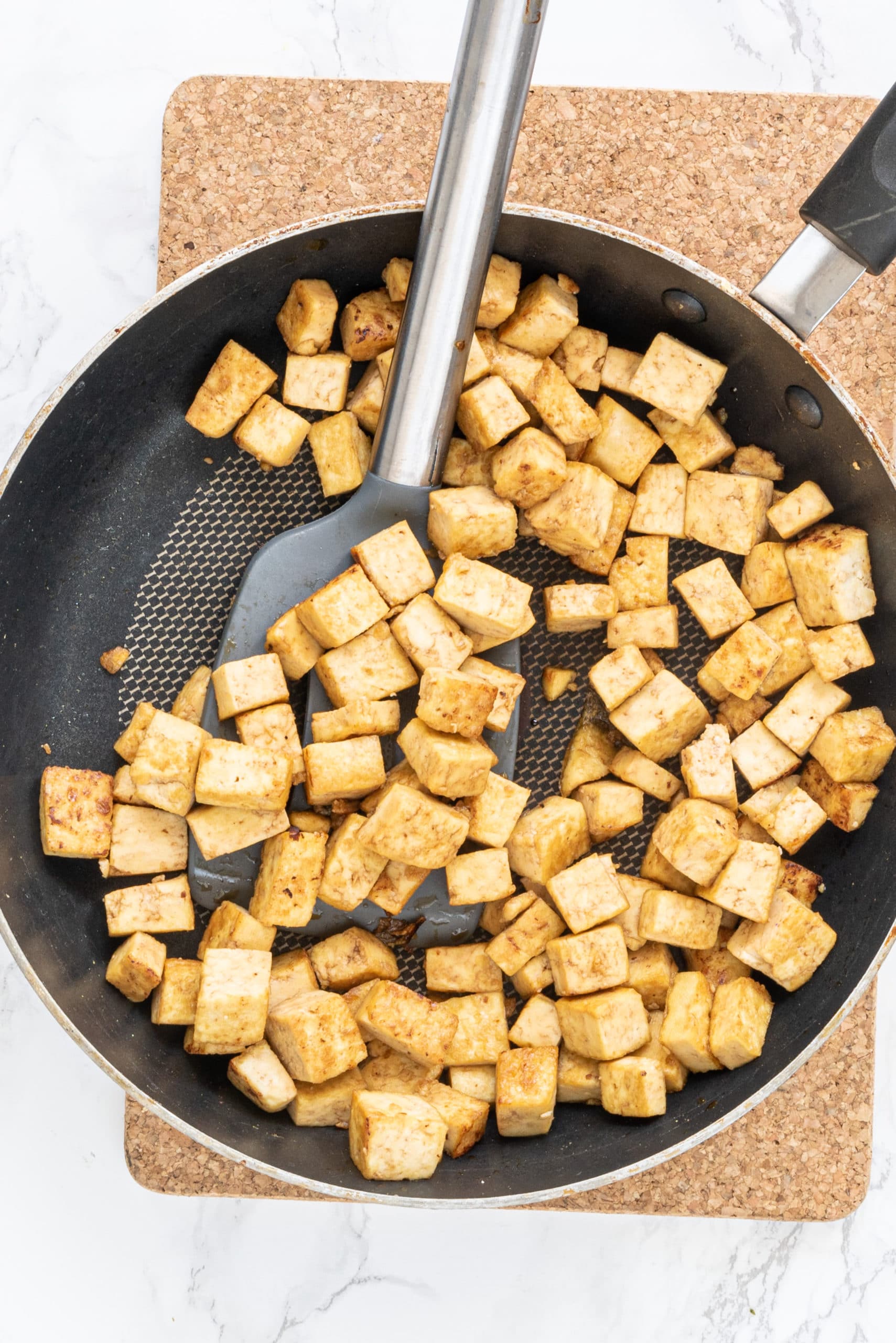 A black frying pan on a cork cutting board filled with tofu cubes and a silicone spatula.