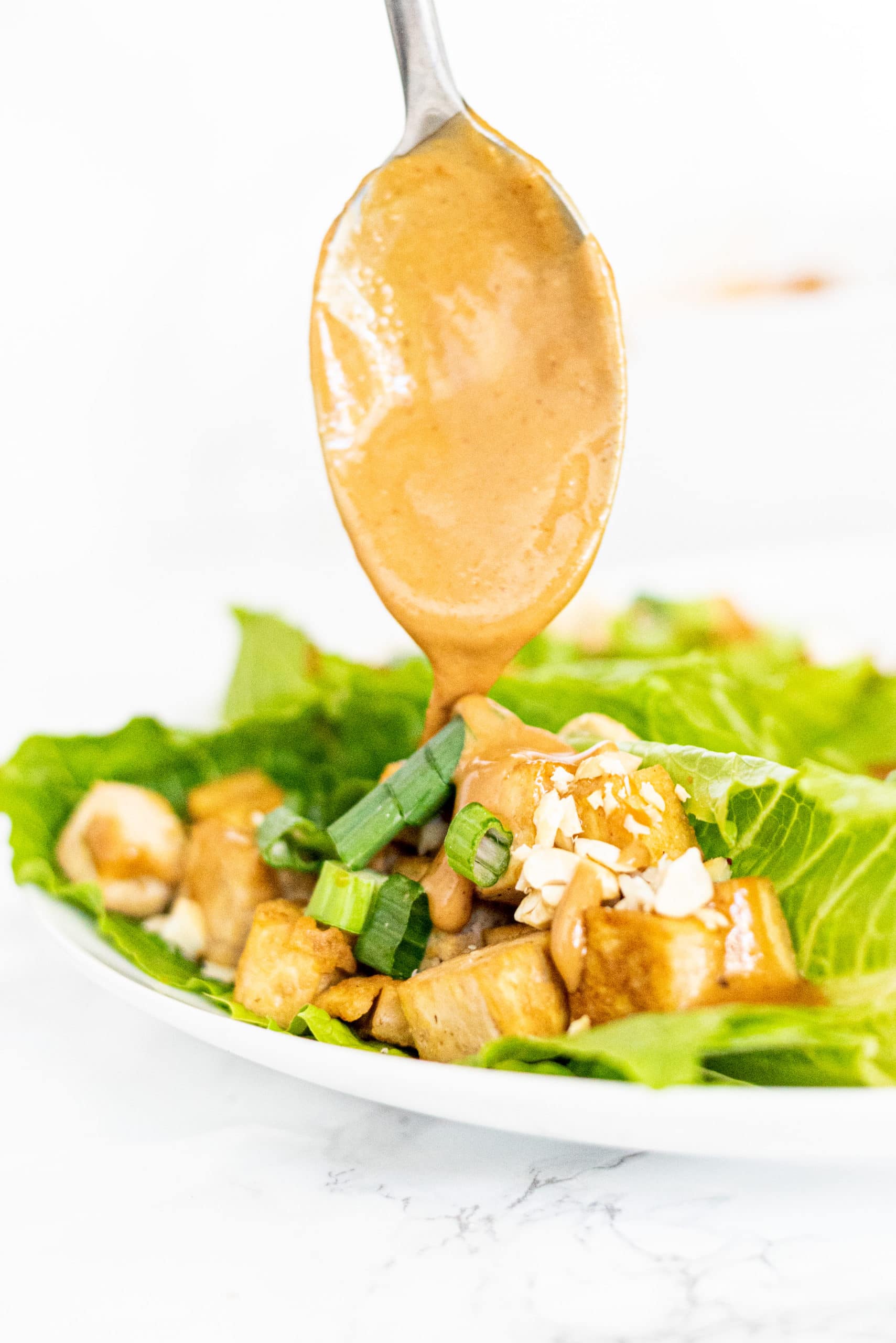 A white plate with lettuce wraps filled with tofu and cashews and green onion and a silver metal spoon covered in peanut sauce drizzling sauce onto the lettuce wraps.