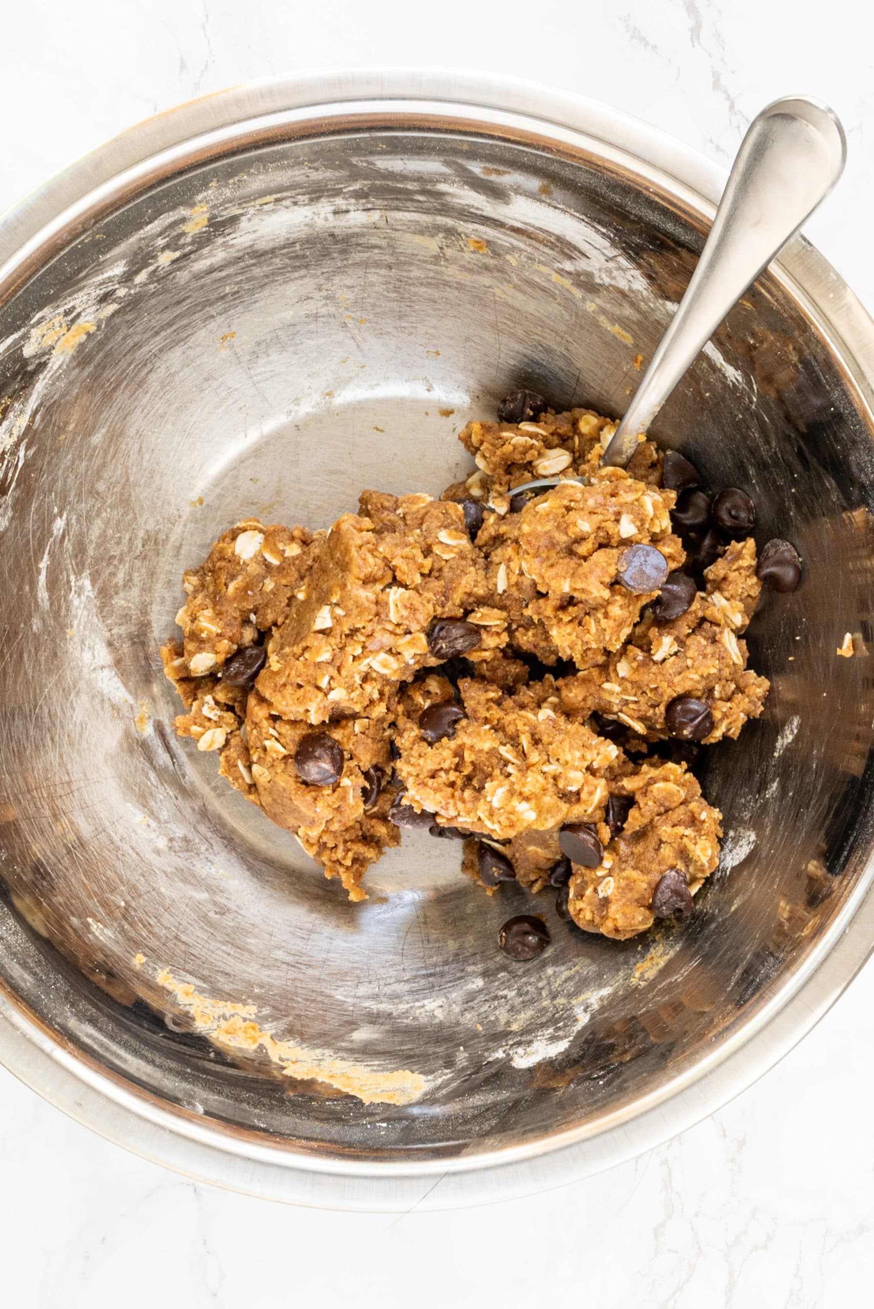 A silver mixing bowl filled with chocolate chip cookie dough and a metal spoon.