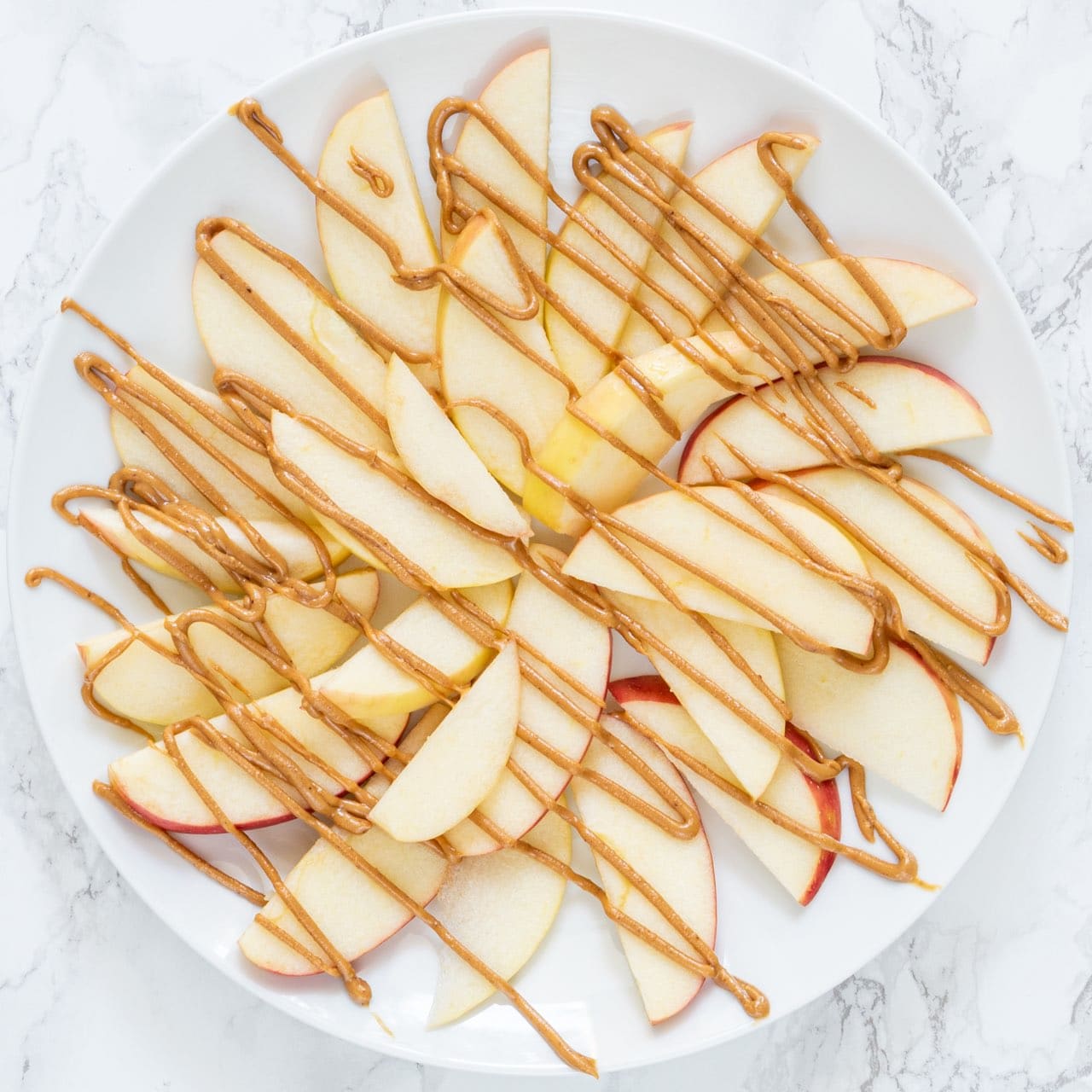 A white plate with apple slices arranged in a flat layer with a drizzle of peanut butter on top.