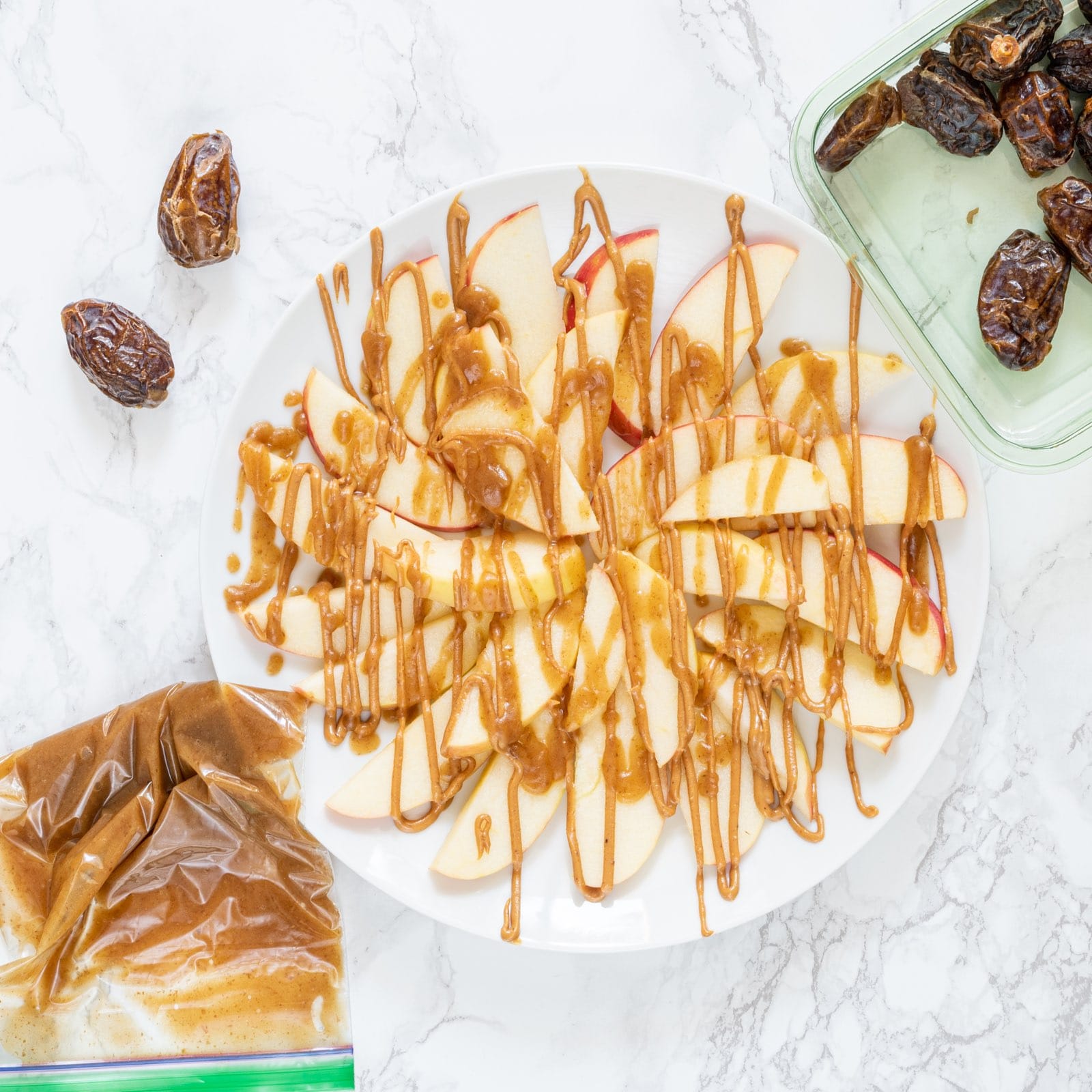 A white plate with apples drizzled with peanut butter on it, and a container of dates.