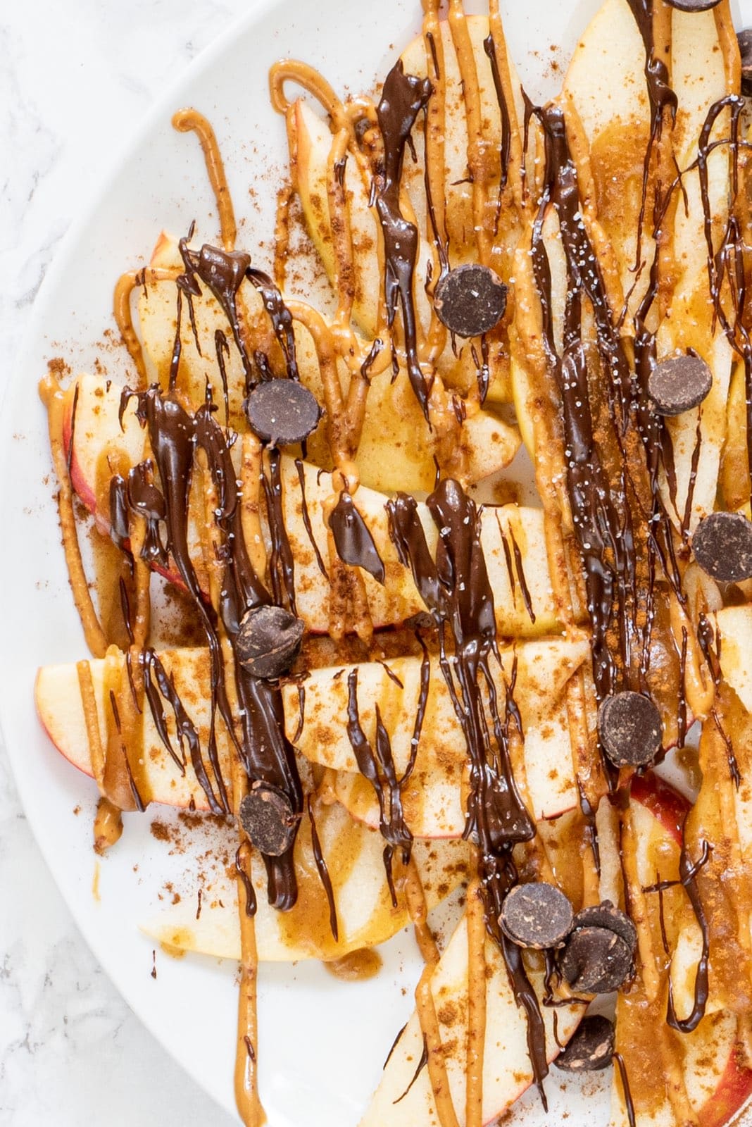 A white plate with apple dessert nachos on it topped with drizzled peanut butter and chocolate.