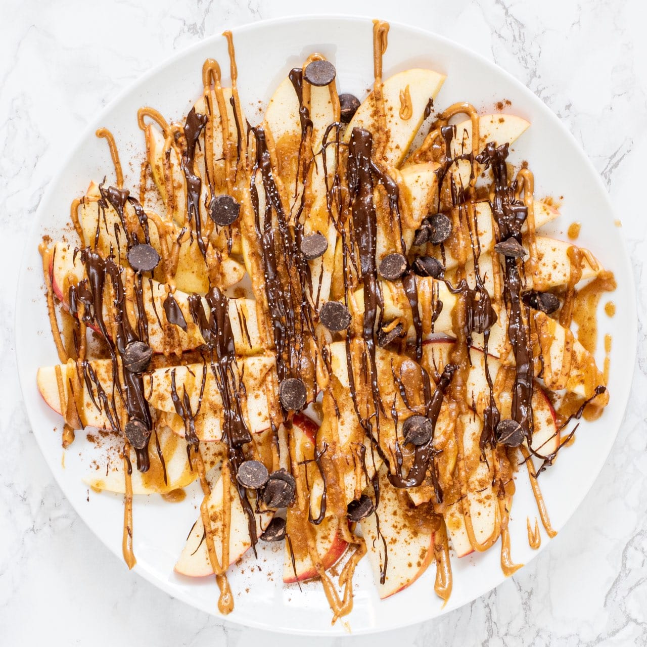 A white plate with apple dessert nachos on it topped with drizzled peanut butter, chocolate, and chocolate chips.