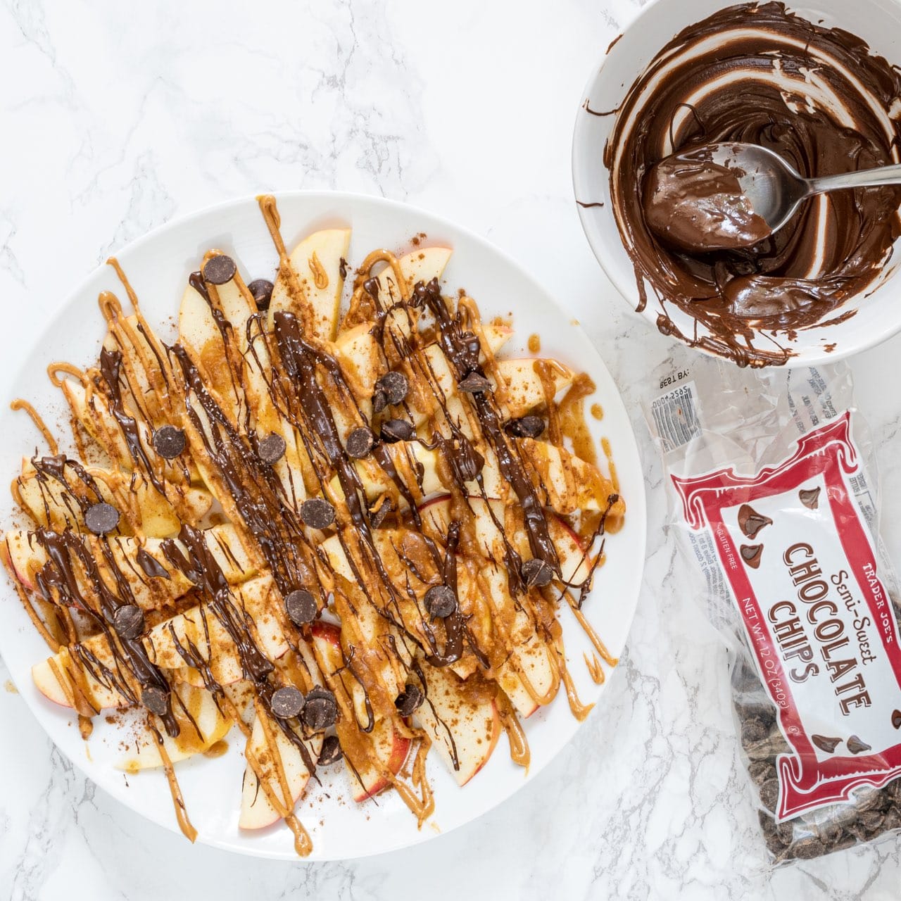 A white plate with sliced apples on it drizzled with peanut butter, chocolate, date caramel, and a small bowl of melted chocolate with a bag of chocolate chips beside it.