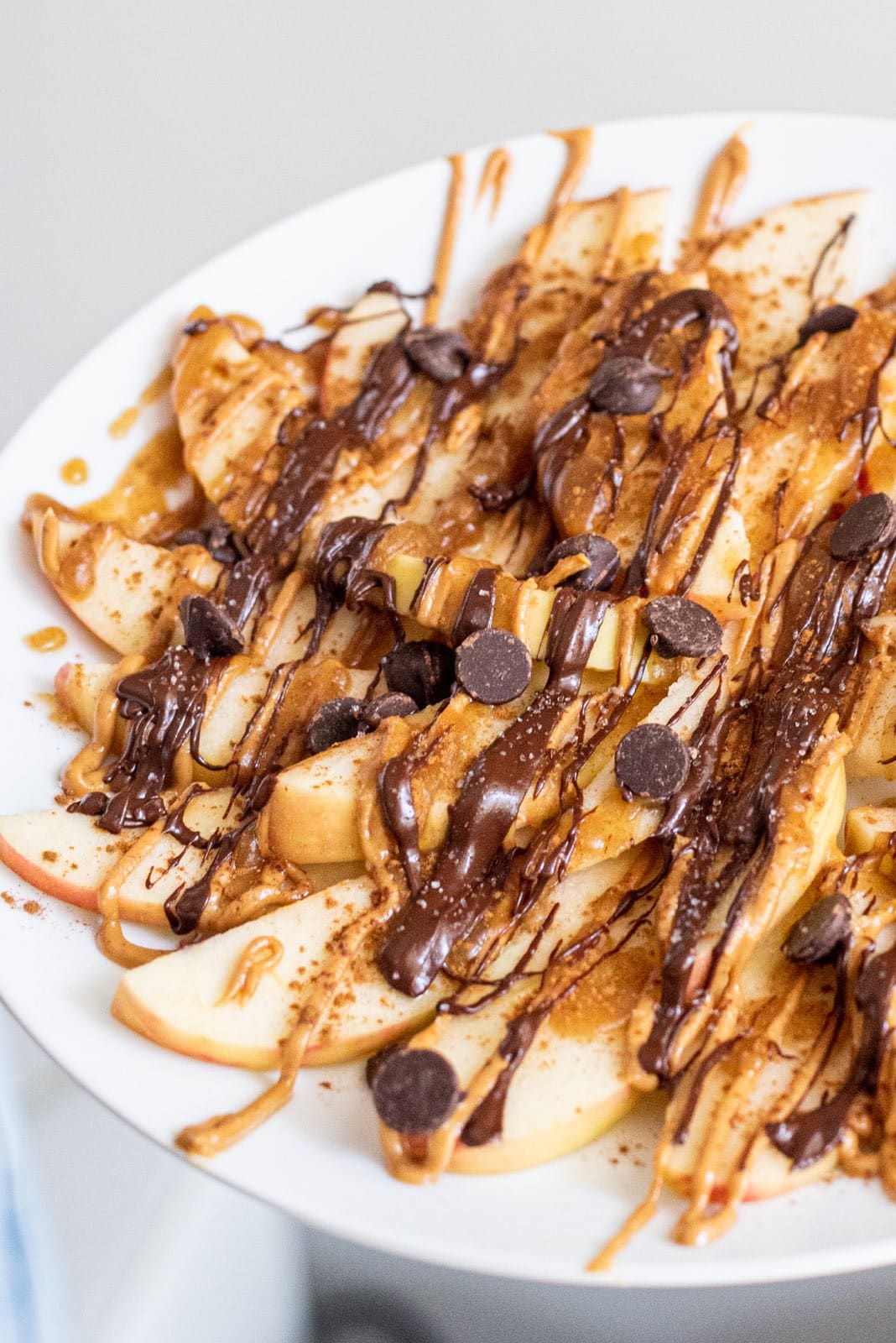 A white plate with apple dessert nachos on it topped with drizzled peanut butter, chocolate, and chocolate chips.