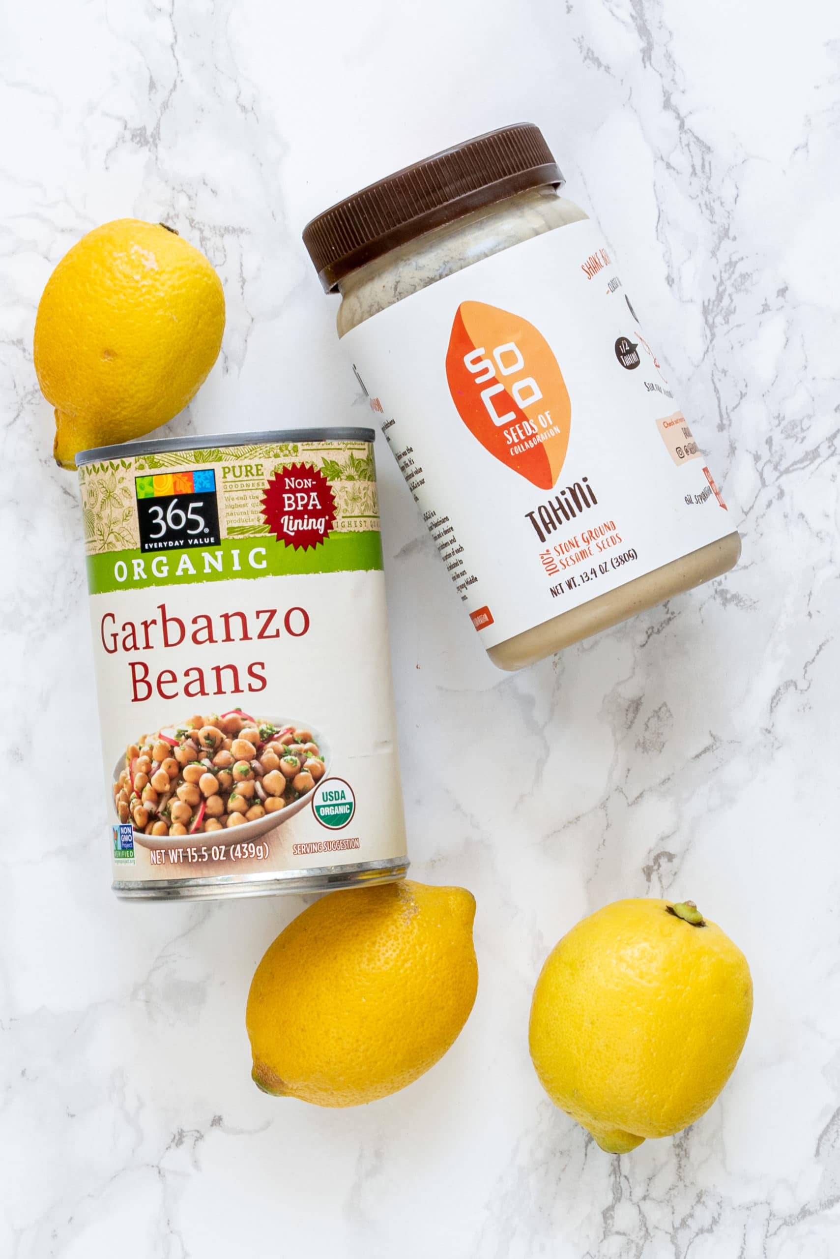 Ingredients laid out on a marble counter to make vegan hummus including: three lemons, tahini, and a can of chickpeas.