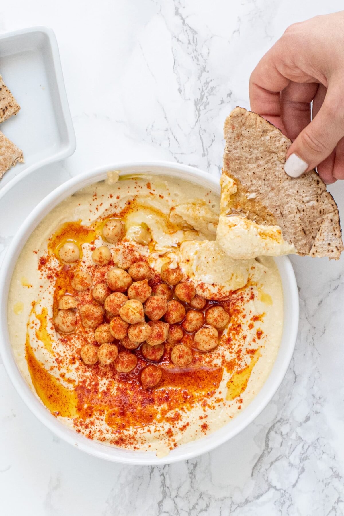 a white bowl filled with vegan hummus topped with chickpeas, paprika, and olive oil. there is a white hand in the right of the photo holding a piece of pita bread dipped into the hummus.