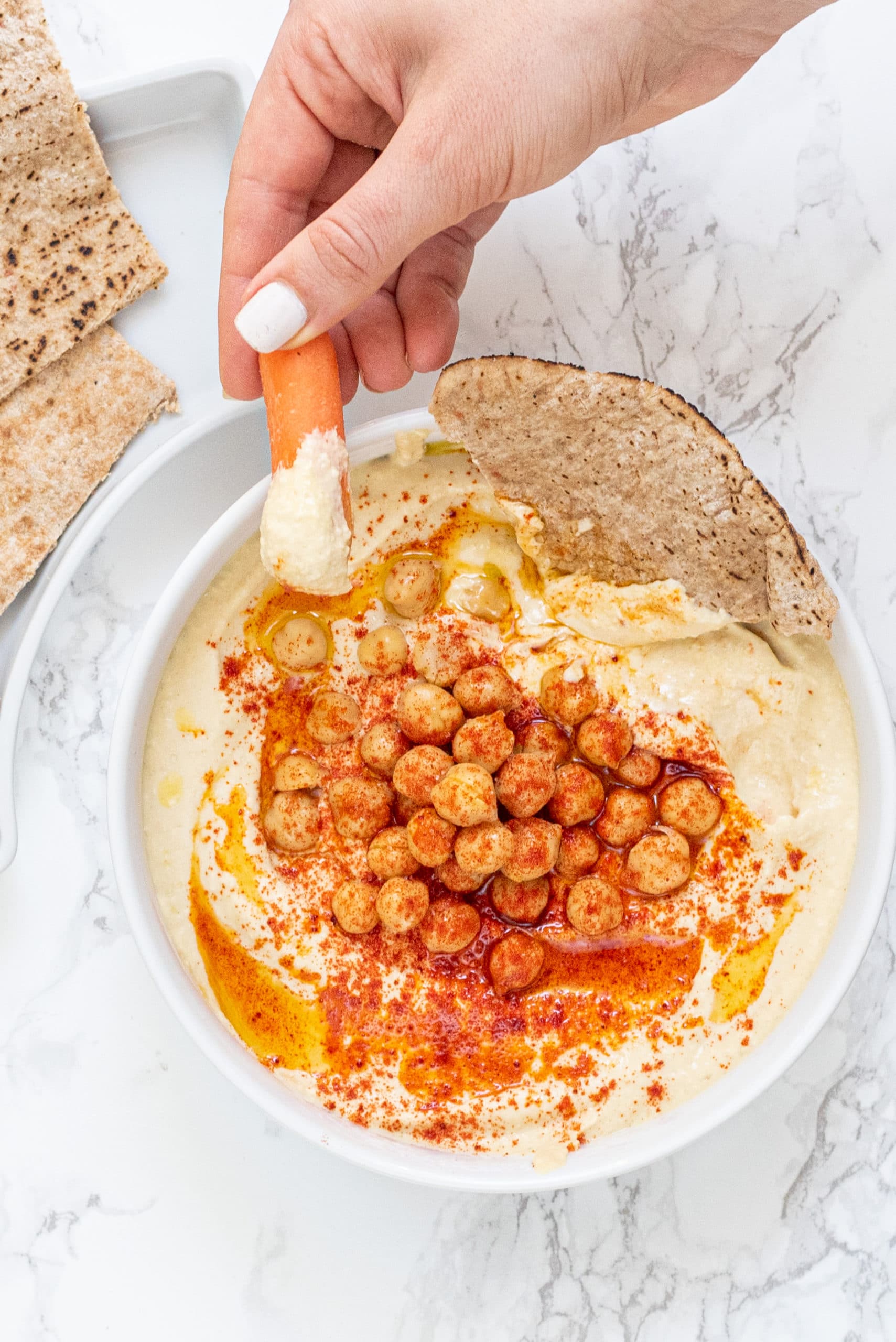 A white bowl filled with hummus and pieces of pita on the side. Plus a white hand coming in from the right side dipping a carrot in the hummus.