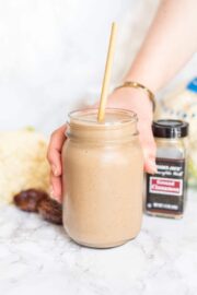 Cinnamon Bun Cauliflower Smoothie in a small mason jar with a straw sticking out and a white hand holding the glass. Behind the smoothie is a cinnamon jar, two medjool dates, and a head of cauliflower.