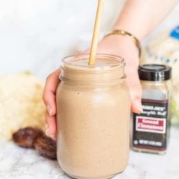 Cinnamon Bun Cauliflower Smoothie in a small mason jar with a straw sticking out and a white hand holding the glass. Behind the smoothie is a cinnamon jar, two medjool dates, and a head of cauliflower.