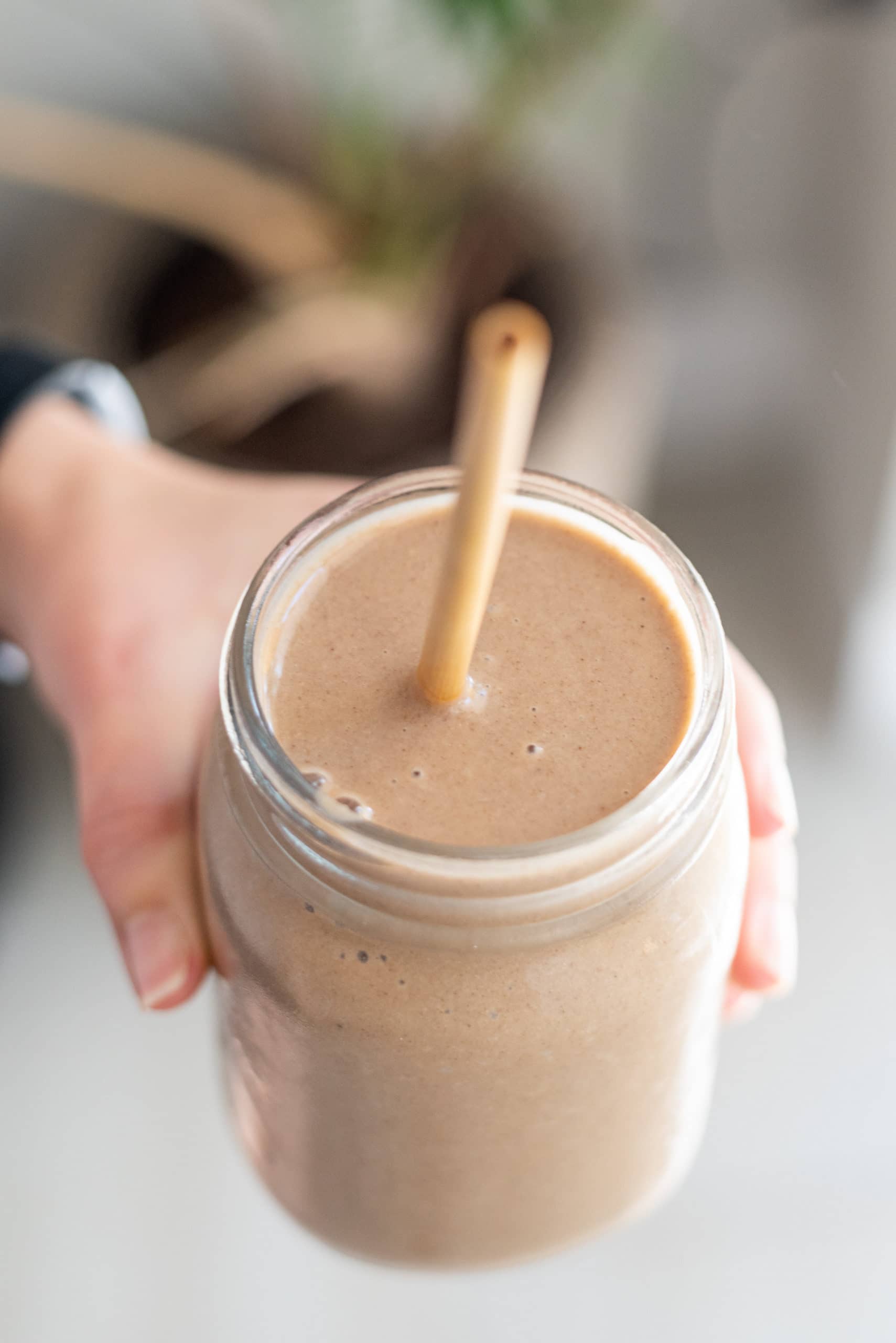 Cinnamon Bun Cauliflower Smoothie in a small glass jar with a straw sticking out. A white hand is holding the smoothie.