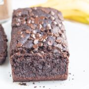 A white plate with a loaf of Vegan Banana Brownie Bread on it with a slice cut off on the end. Behind the plate is two bananas and a jar of cacao powder.