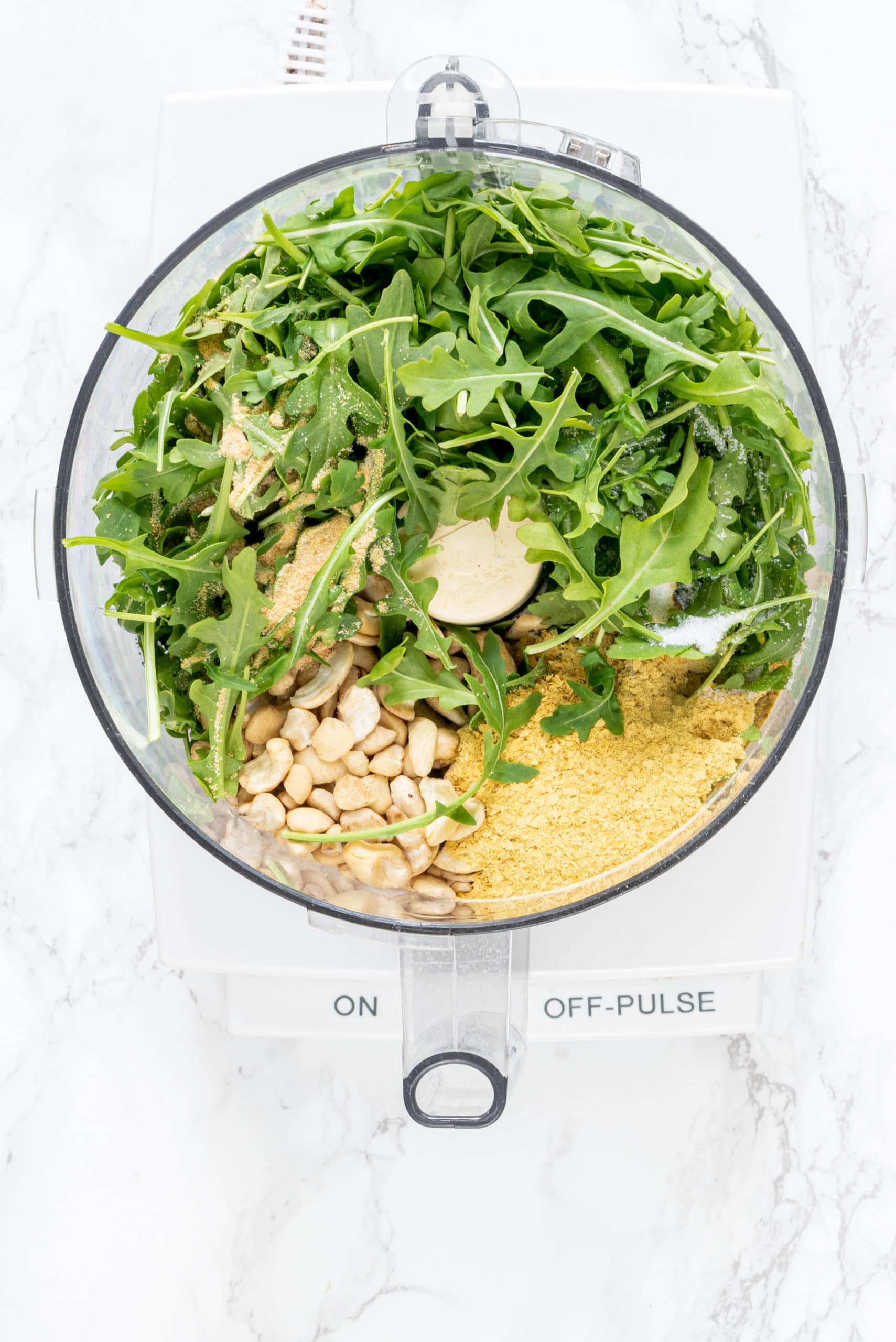A food processor filled with cashews, nutritional yeast, arugula, and basil.