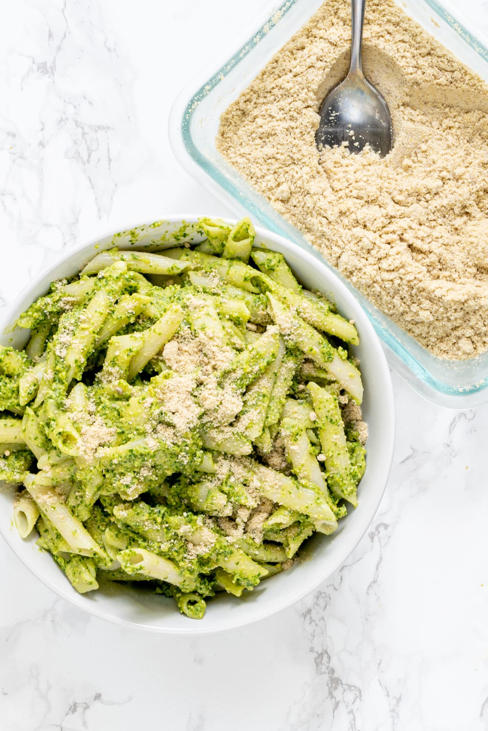 A bowl of pesto penne pasta topped with a sprinkle of parmesan cheese and a clear glass dish beside the bowl filled with parmesan cheese and a silver metal spoon in it.