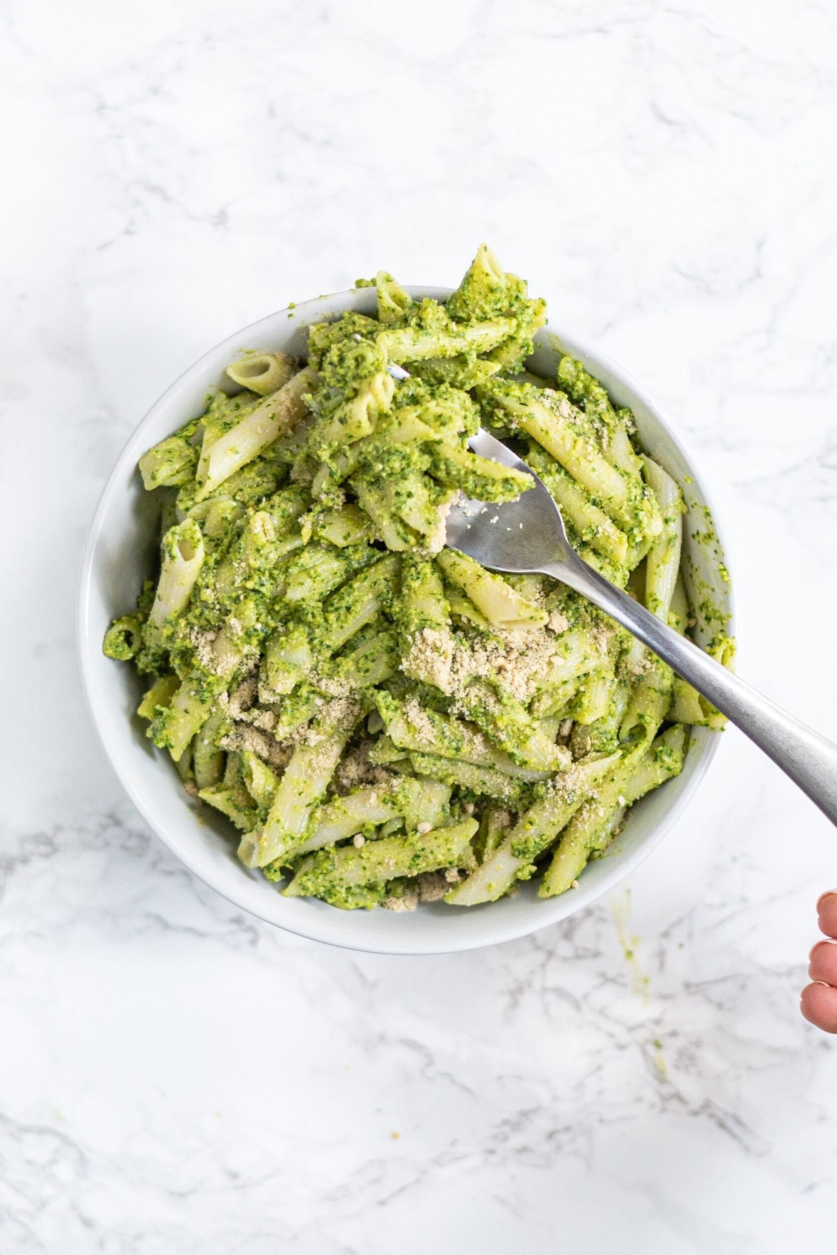 A white bowl filled with pesto pasta and a silver metal fork in the bowl.