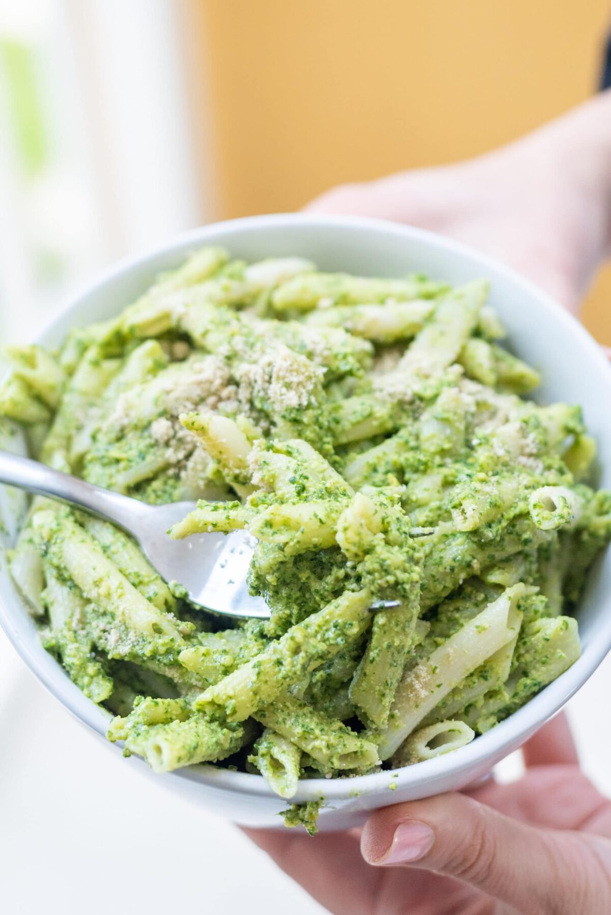 Two white hands holding a white bowl filled with pesto penne pasta with a fork in it and sprinkled cheese on top.