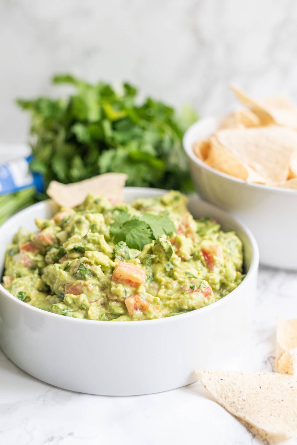 A white bowl of guacamole topped with cilantro. Beside the bowl is a bowl of chips, fresh limes, and fresh cilantro.