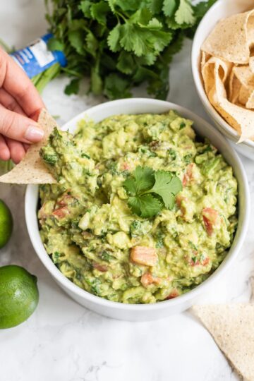 A white bowl of guacamole topped with cilantro. Beside the bowl is a bowl of chips, fresh limes, and fresh cilantro.