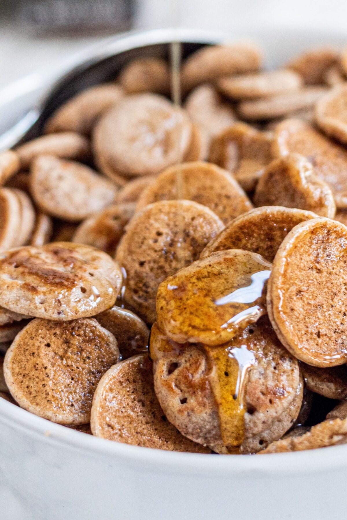 A white bowl of pancake cereal topped with maple syrup and a spoon in the bowl.