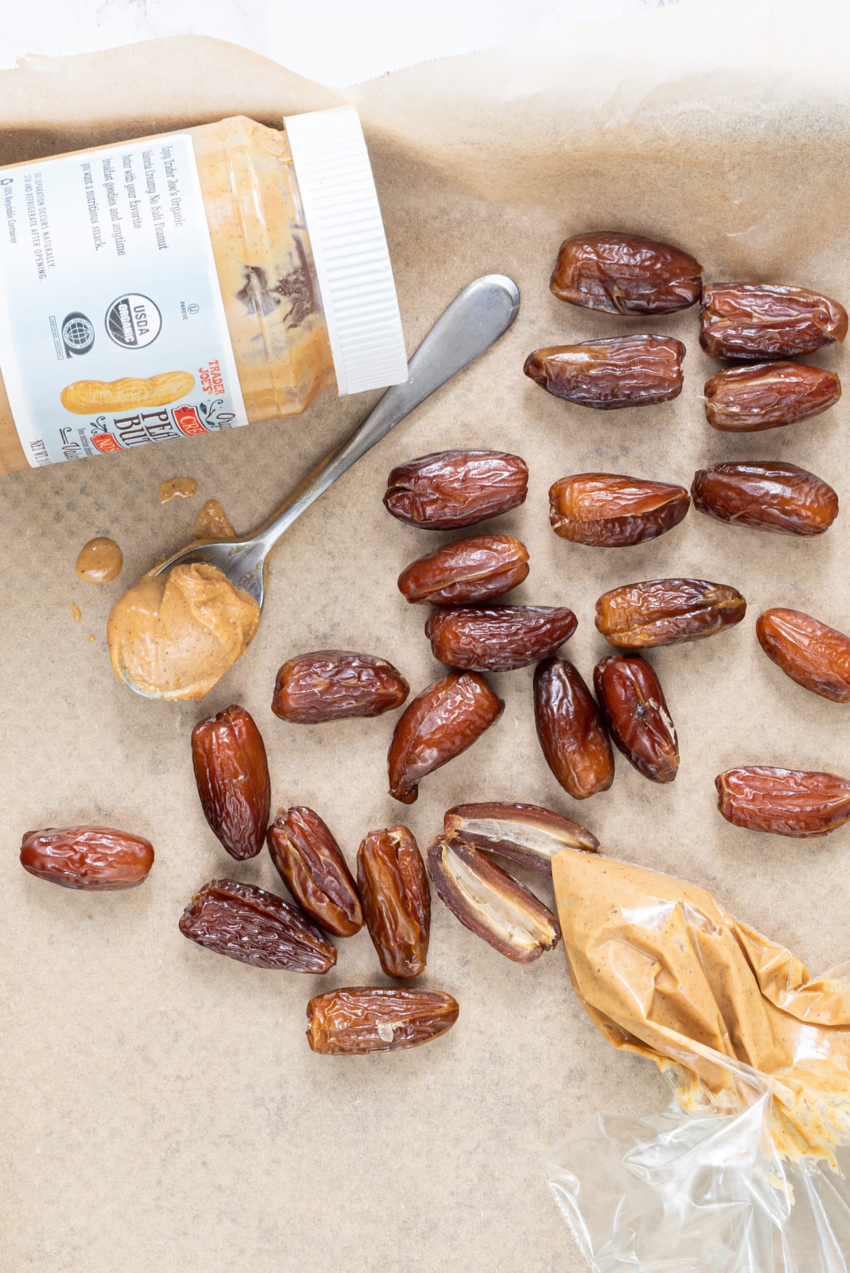 A parchment lined baking sheet with dates on it, a jar of peanut butter, a spoon with peanut butter, and a baggie filled with peanut butter.