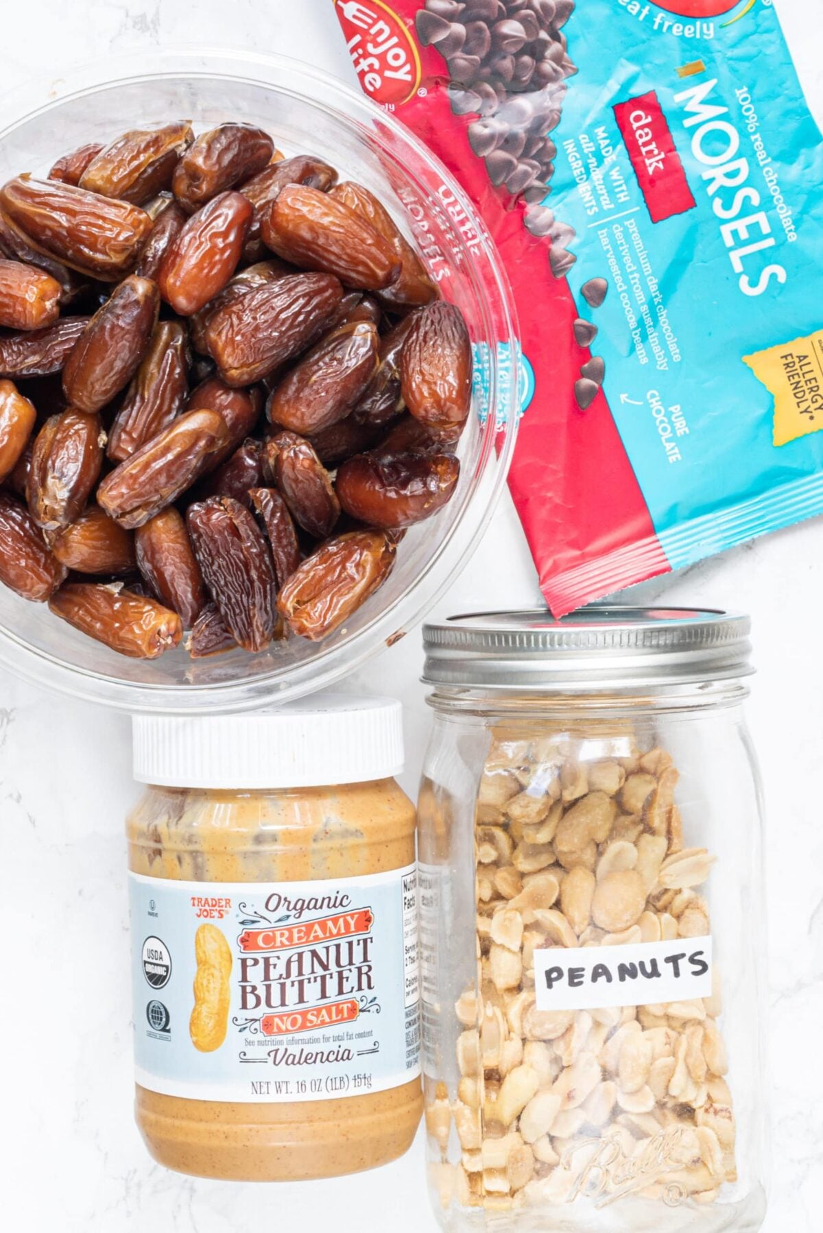 A bowl of dates, a jar of peanut butter, a jar of peanuts, and a bag of chocolate chips.