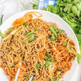 A white bowl filled with vegan pad thai and a set of chopsticks sticking out from the bowl and a bundle of fresh cilantro.