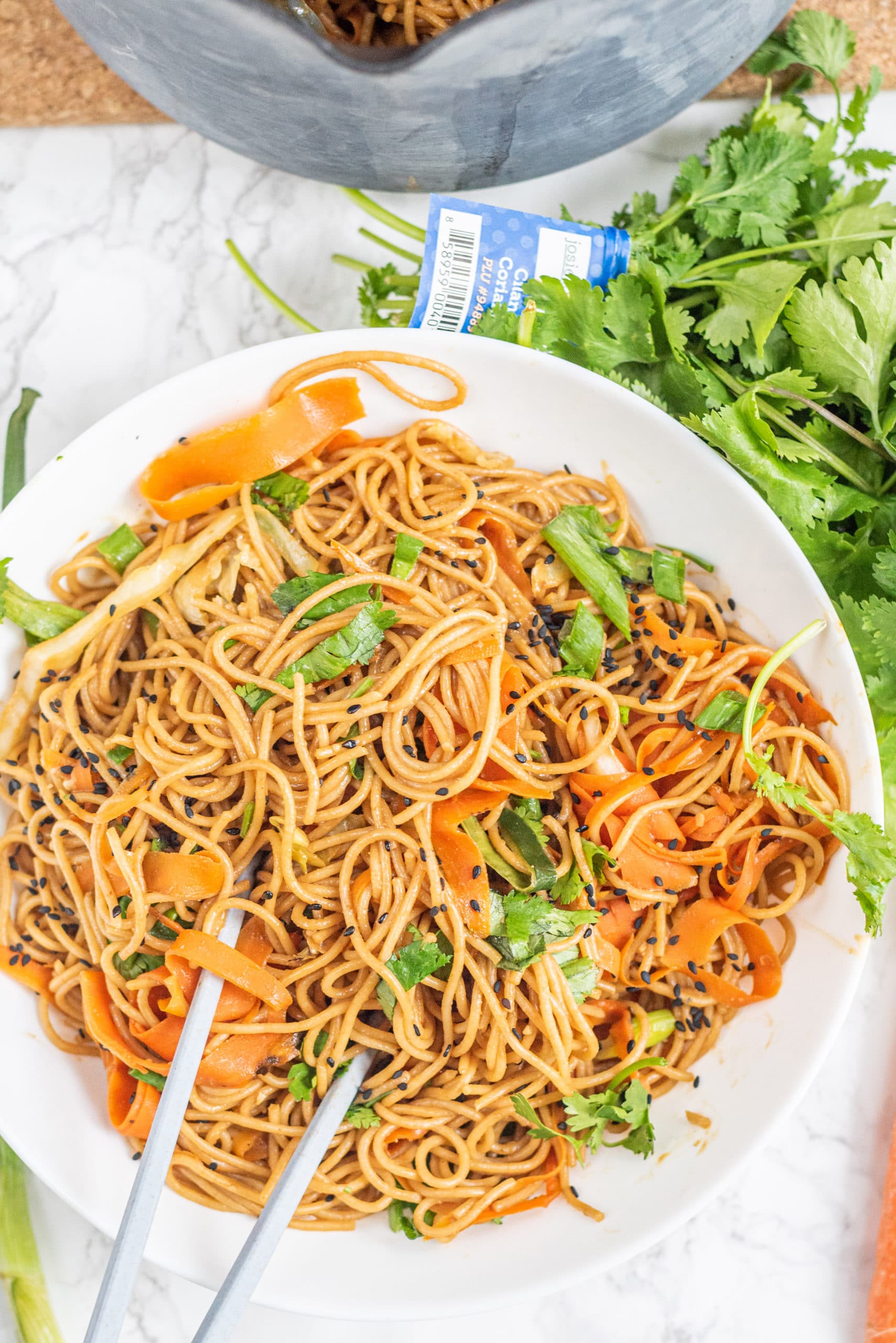 A white bowl filled with vegan pad thai and a set of chopsticks sticking out from the bowl and a bundle of fresh cilantro.