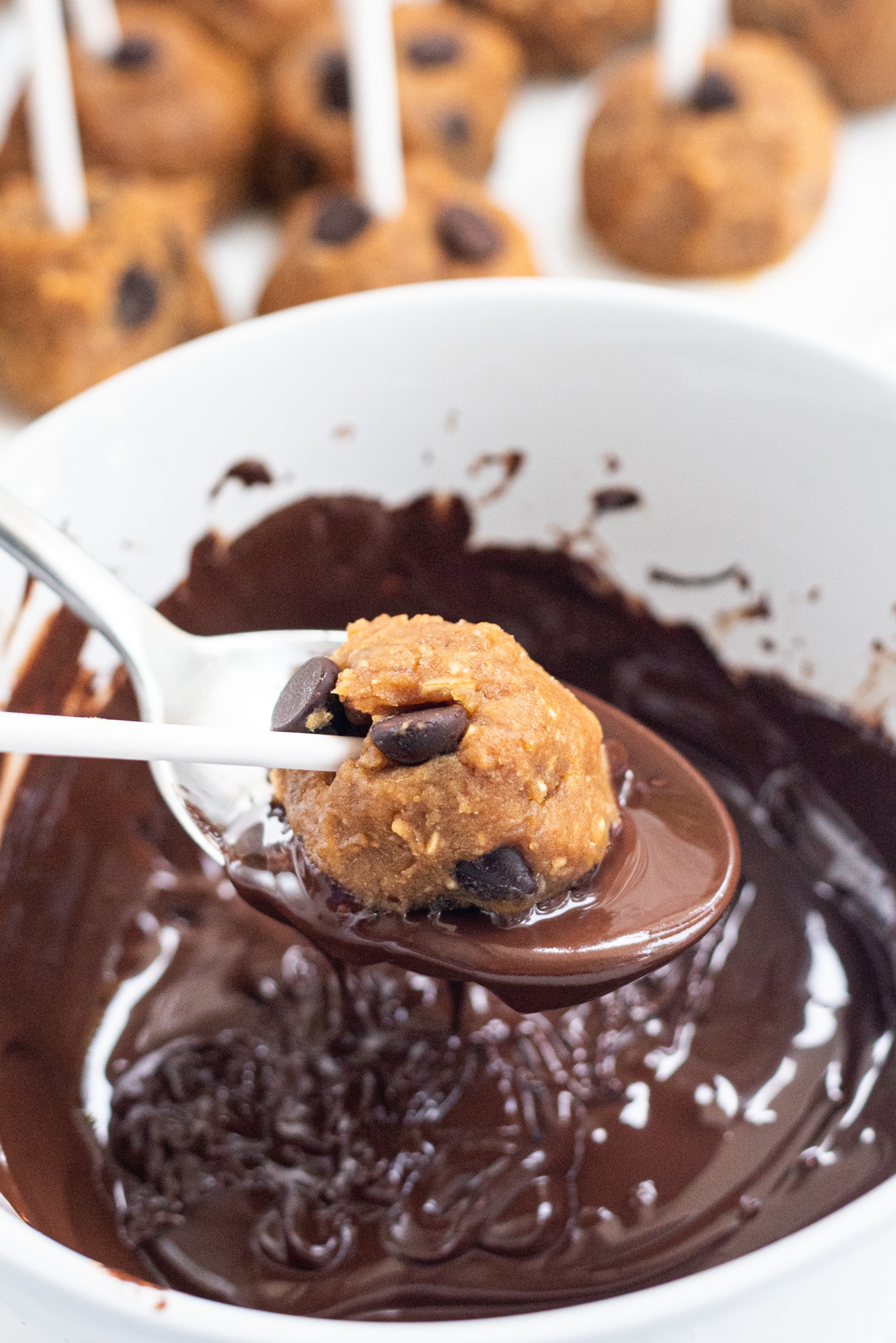 A white bowl of melted chocolate and a spoon with a cookie dough ball in it being rolled in chocolate.