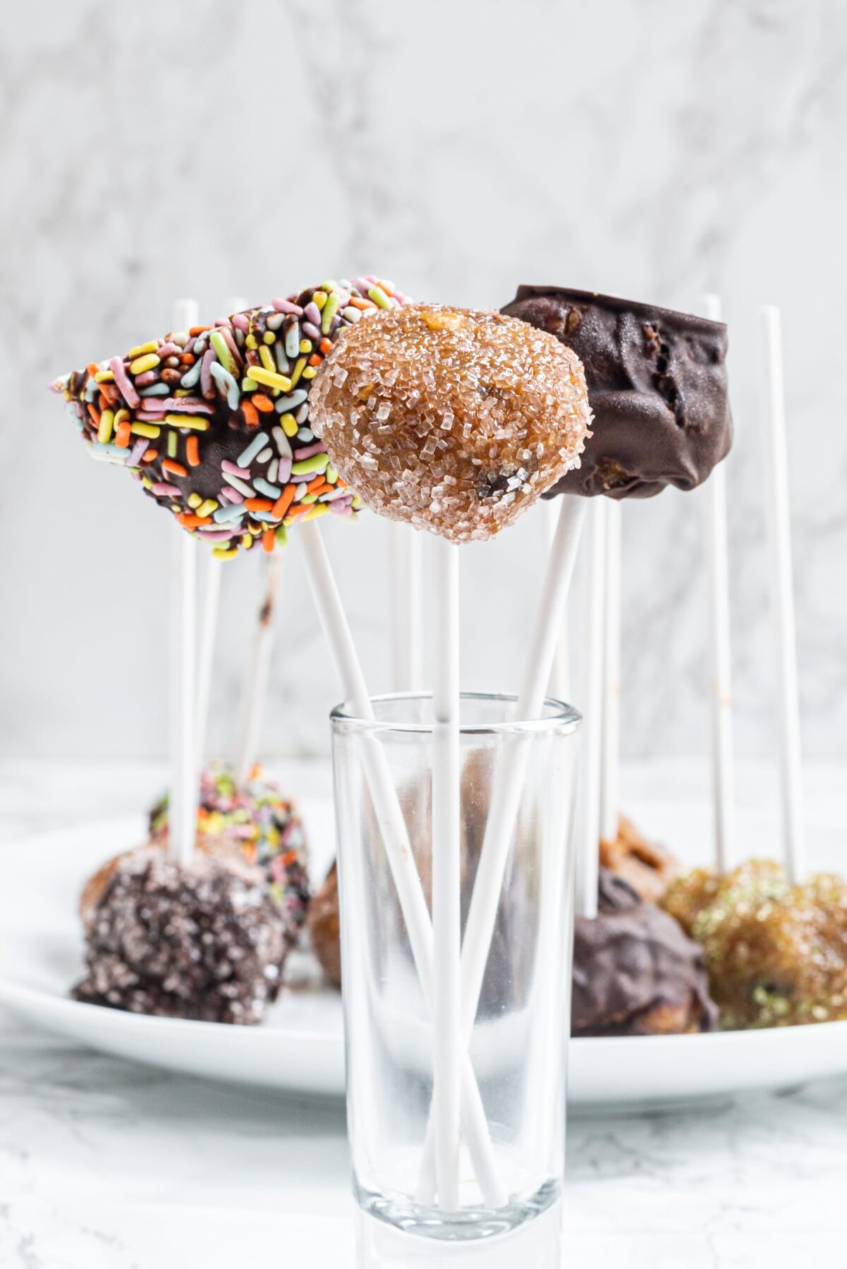 A glass cup holding three cookie dough pops on sticks. One covered with chocolate, one covered with sugar, and one covered with chocolate and sprinkles.