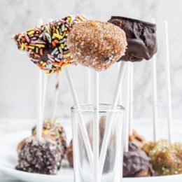 A glass cup holding three cookie dough pops on sticks. One covered with chocolate, one covered with sugar, and one covered with chocolate and sprinkles.