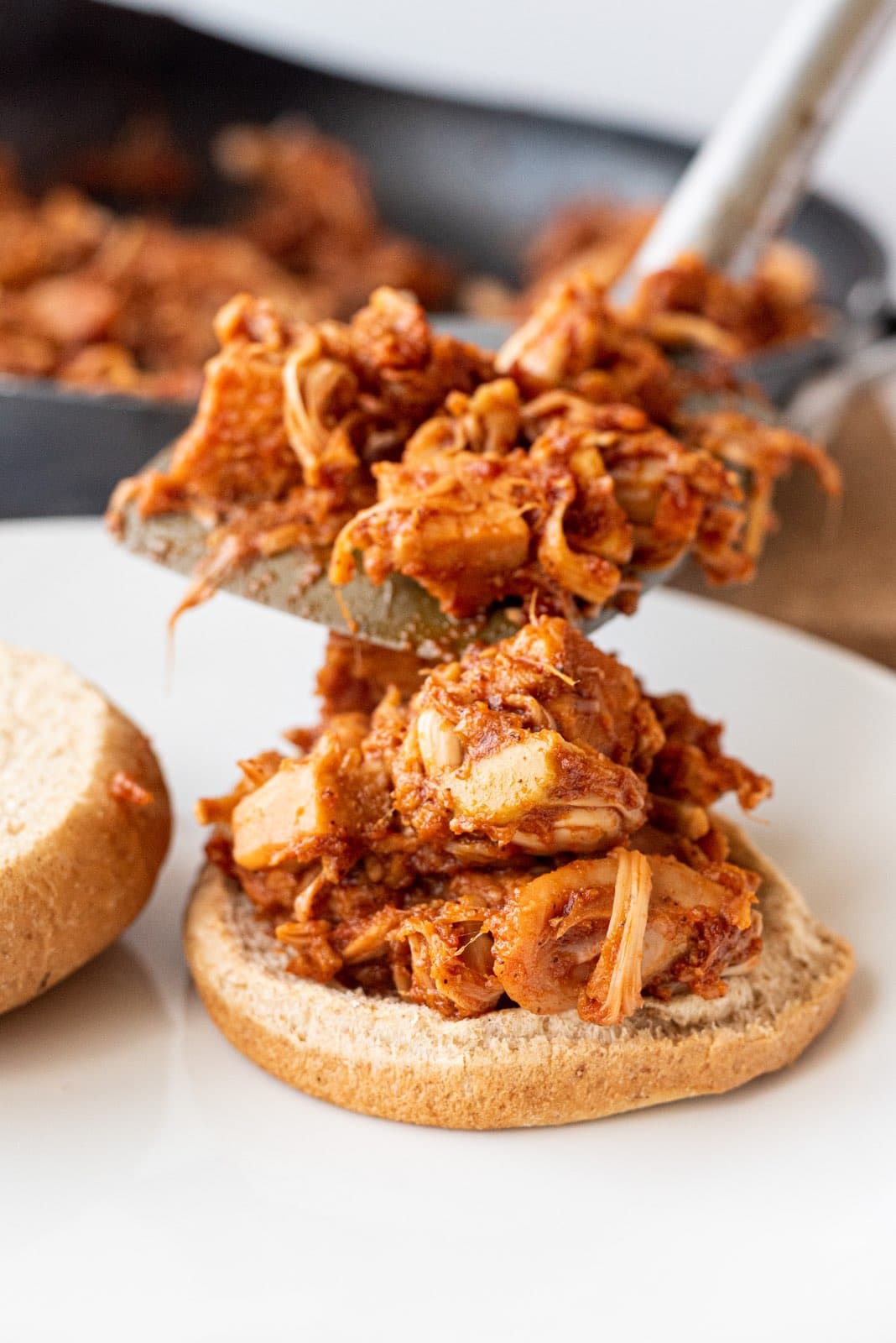 A bottom burger bun topped with BBQ jackfruit on a white plate and a spatula holding more shredded jackfruit.