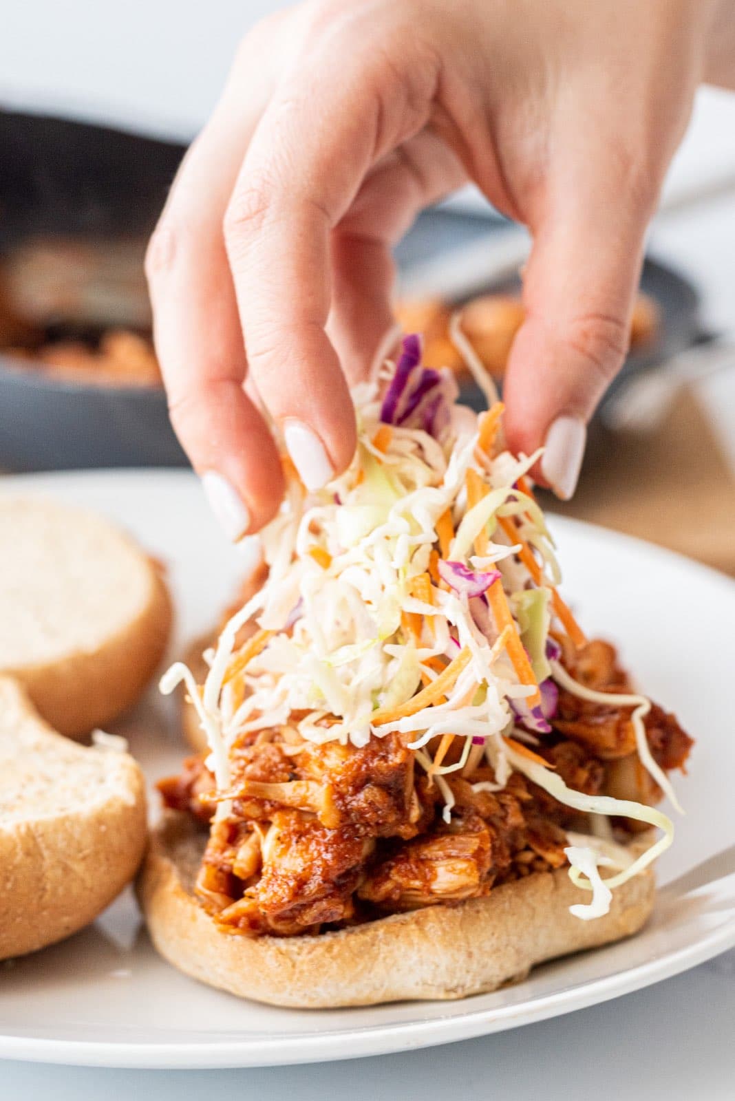 A white hand putting coleslaw on top of pulled jackfruit on a burger bun.