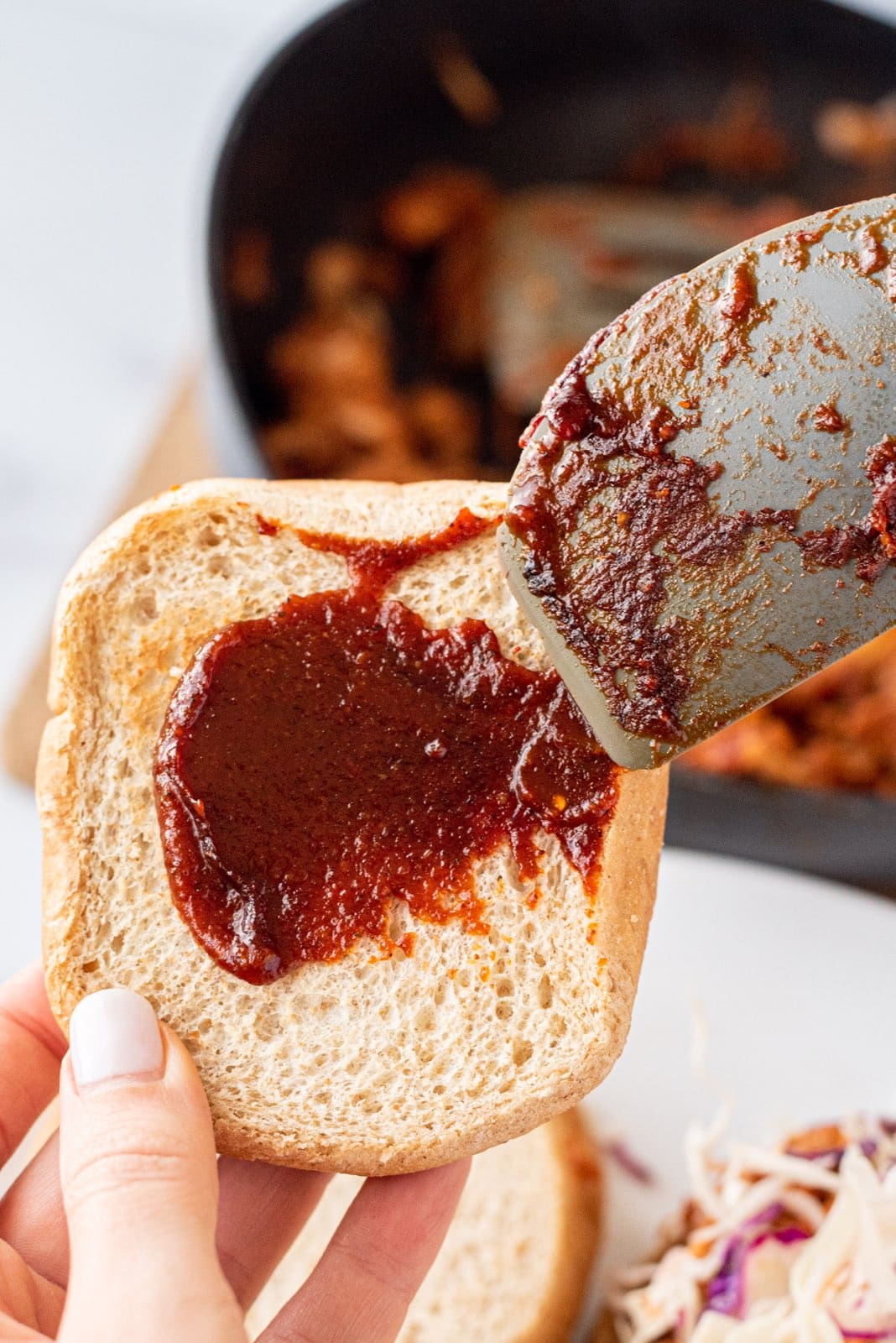 A white hand holding a bun and a spatula spreading BBQ sauce on the bun.
