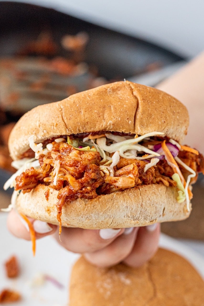 A white hand holding a BBQ jackfruit sandwich on a burger bun with cabbage mix on it.