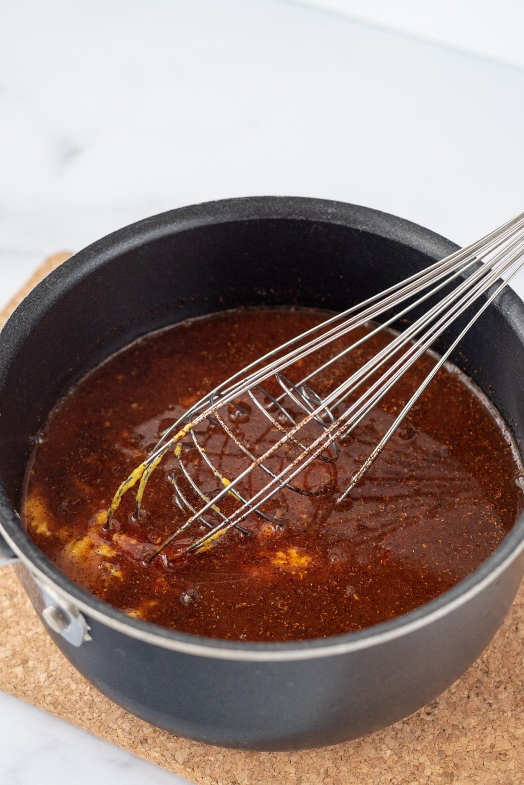 A black saucepan filled with homemade bbq sauce and a silver whisk.
