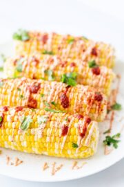 A plate with four ears of corn on the cob on it drizzled with hot sauce, spicy cream sauce, and fresh cilantro.
