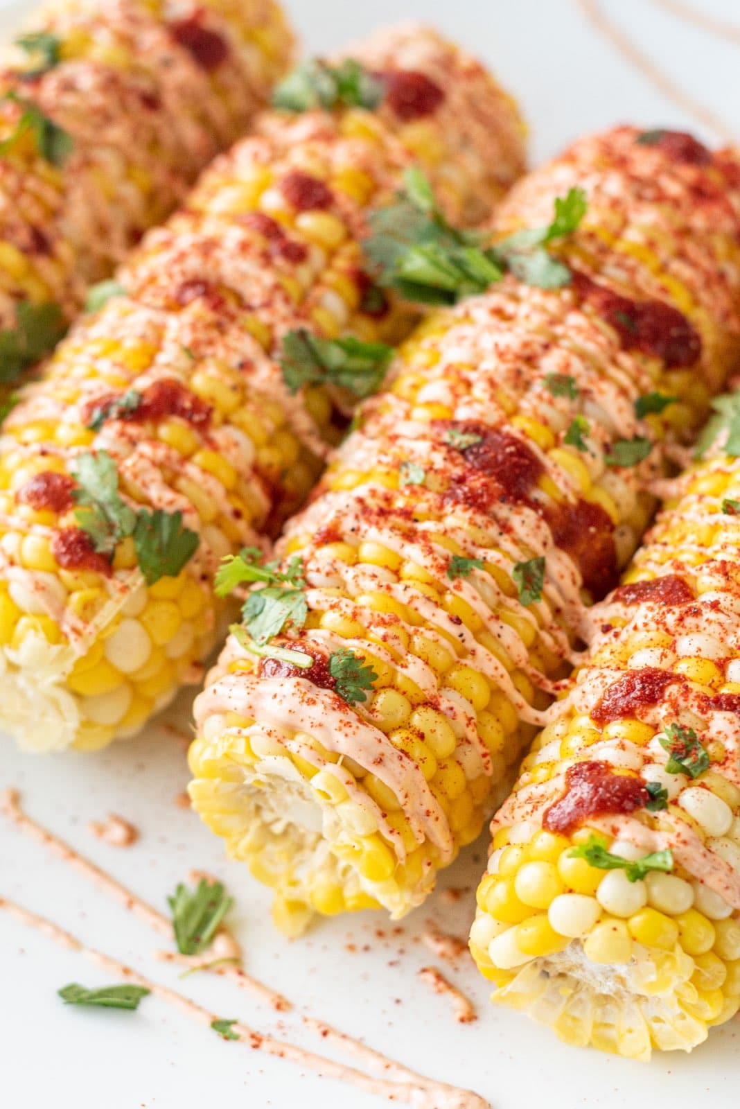 A plate with four ears of corn on the cob on it drizzled with hot sauce, spicy cream sauce, and fresh cilantro.