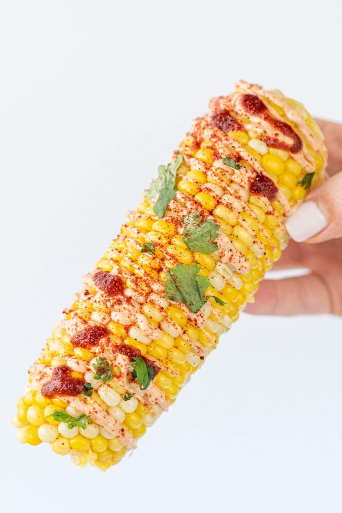 A white hand holding a cob of corn topped with hot sauce and fresh cilantro.